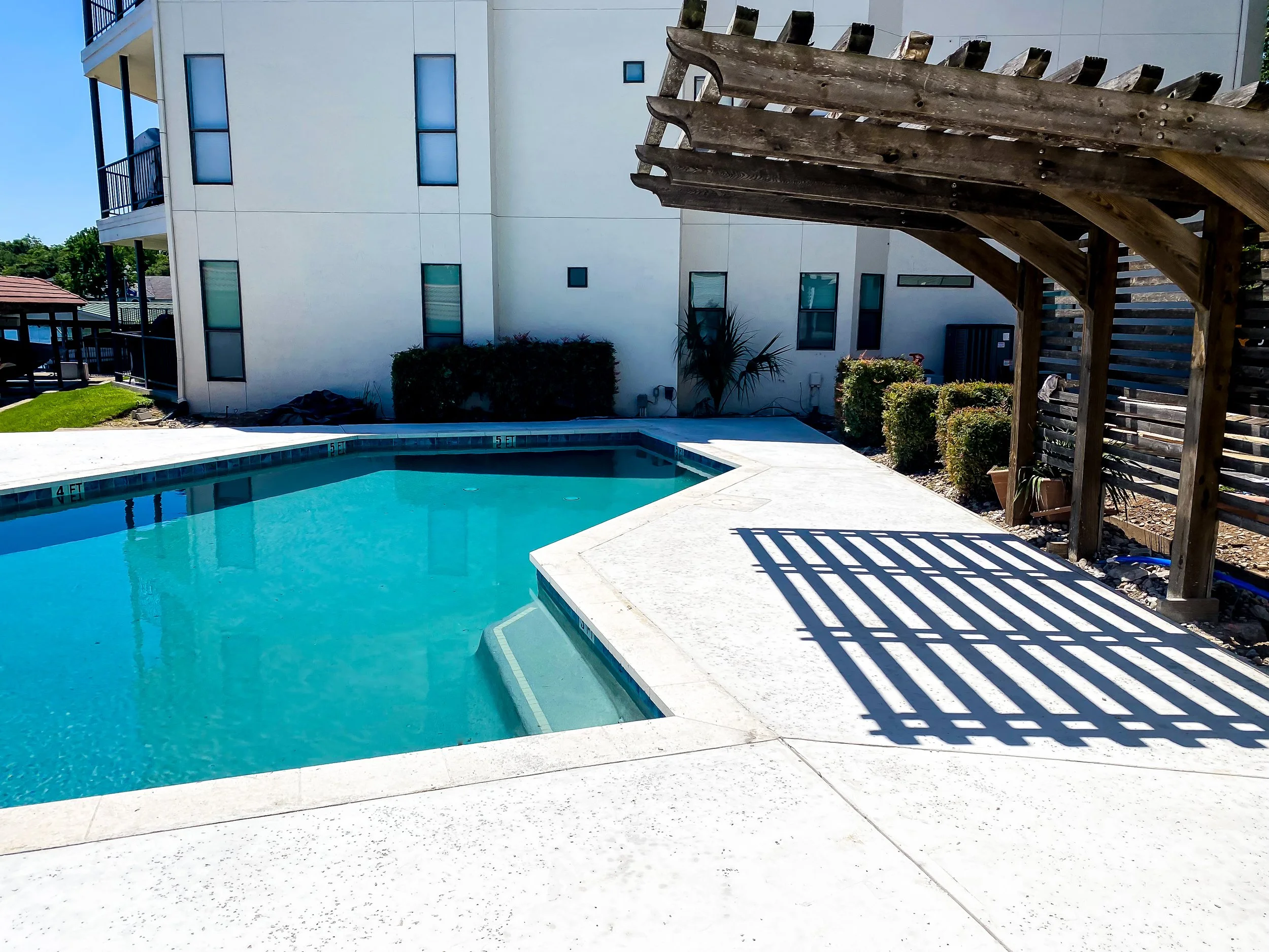 A backyard swimming pool with stairs, surrounded by a concrete deck and a wooden pergola, against a modern white apartment building with small windows and bushes.
