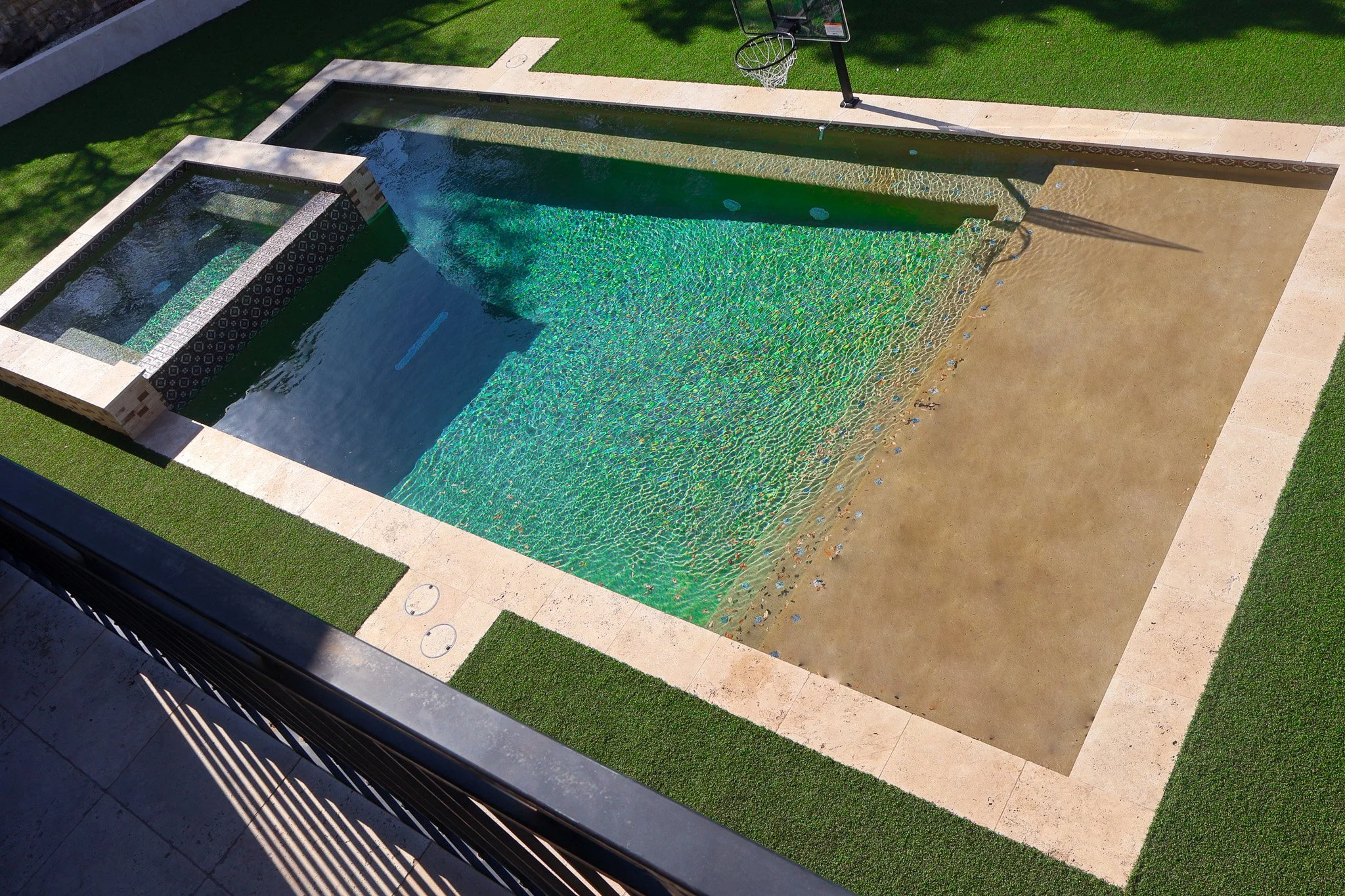 A rectangular swimming pool with a built-in hot tub, with water cascading into the main pool. The pool is surrounded by concrete, grass patches, and a sports hoop on a paved court nearby.