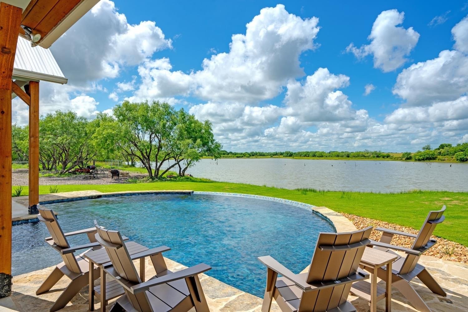 A backyard with a kidney-shaped swimming pool, four Adirondack chairs on a stone patio, a grassy yard, and a large lake with trees on the far side, under a partly cloudy sky.