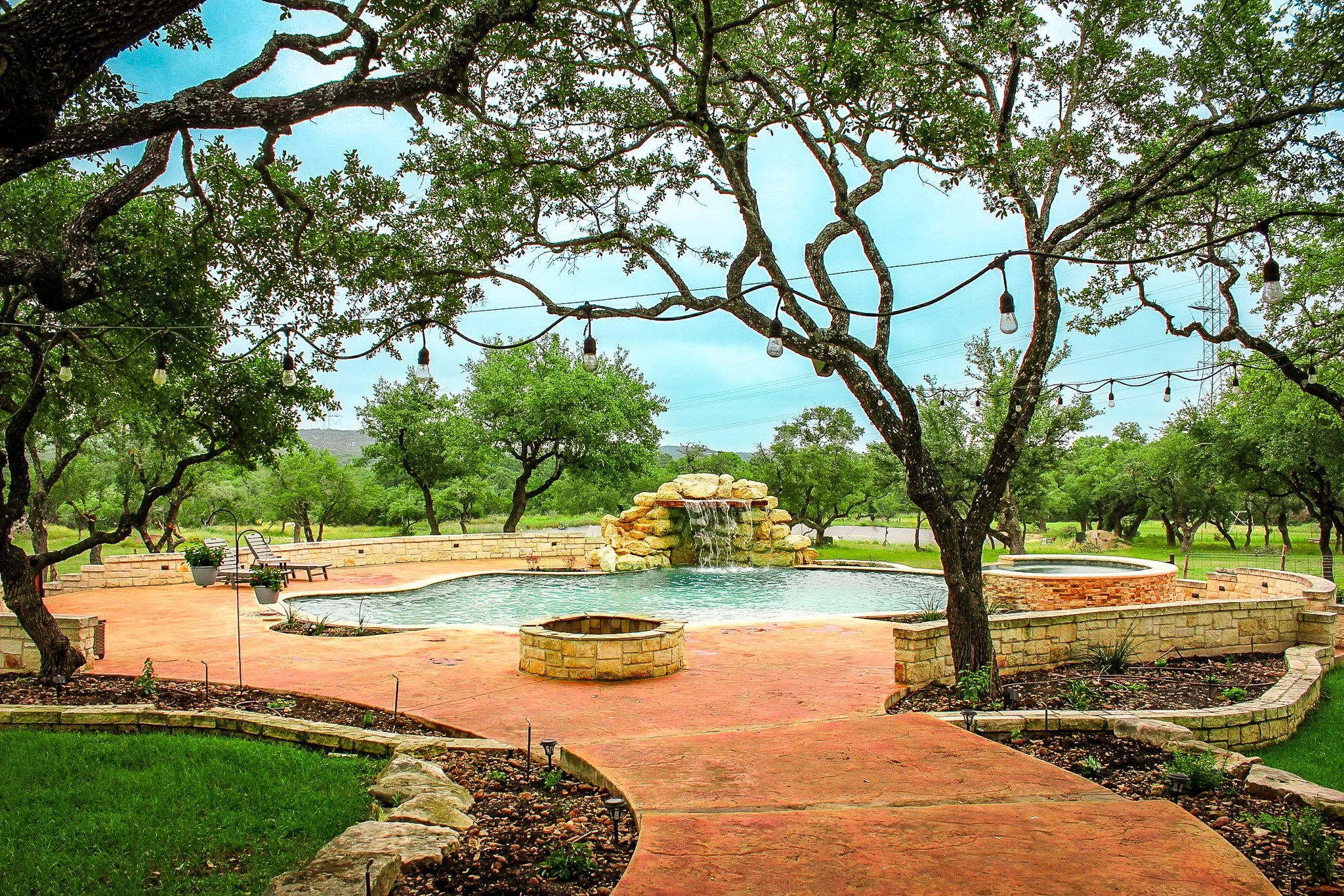 A backyard with a swimming pool, surrounded by trees, string lights, and a stone pathway.