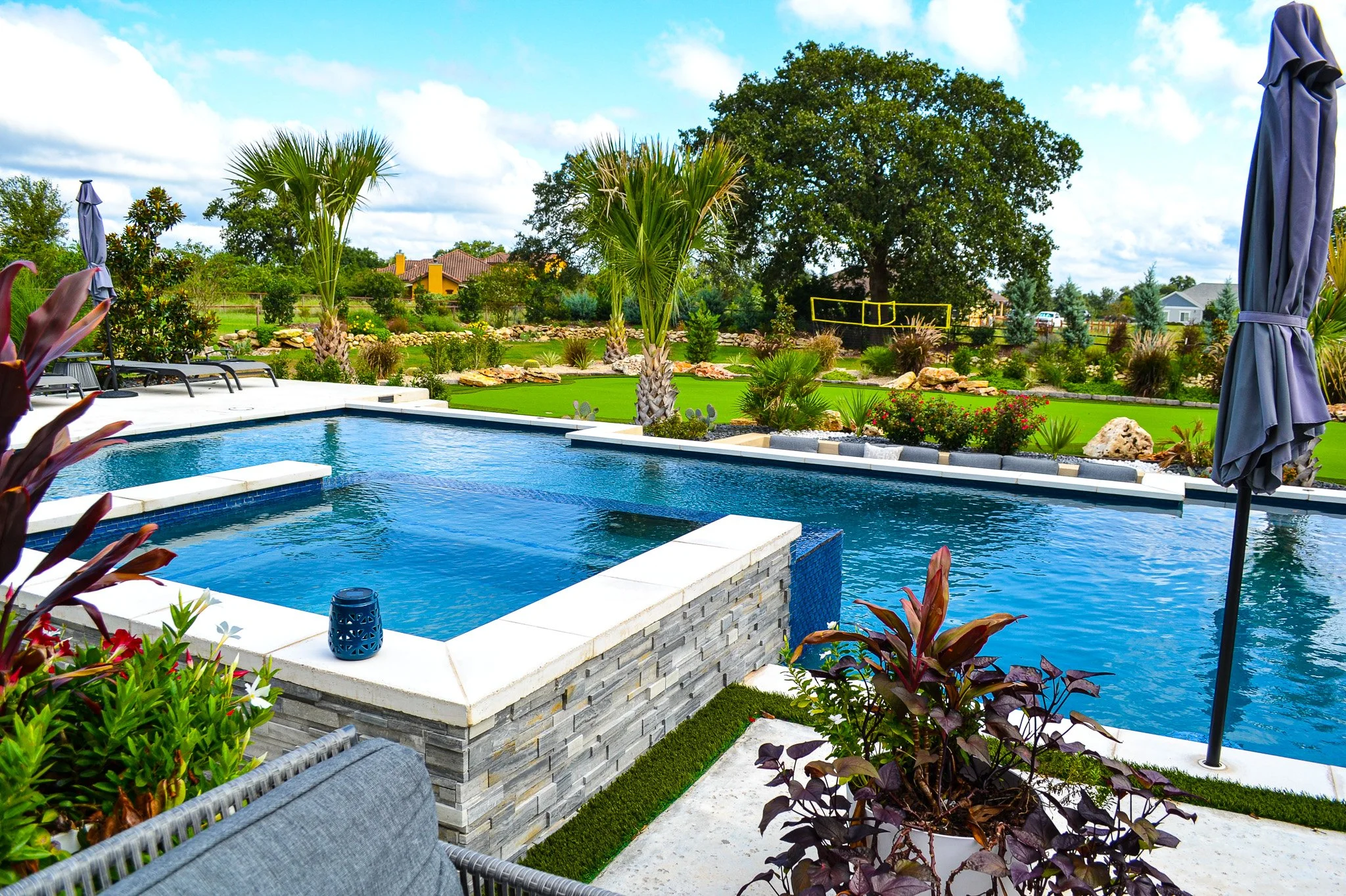 Backyard swimming pool surrounded by plants, trees, and outdoor furniture on a sunny day with partly cloudy sky.
