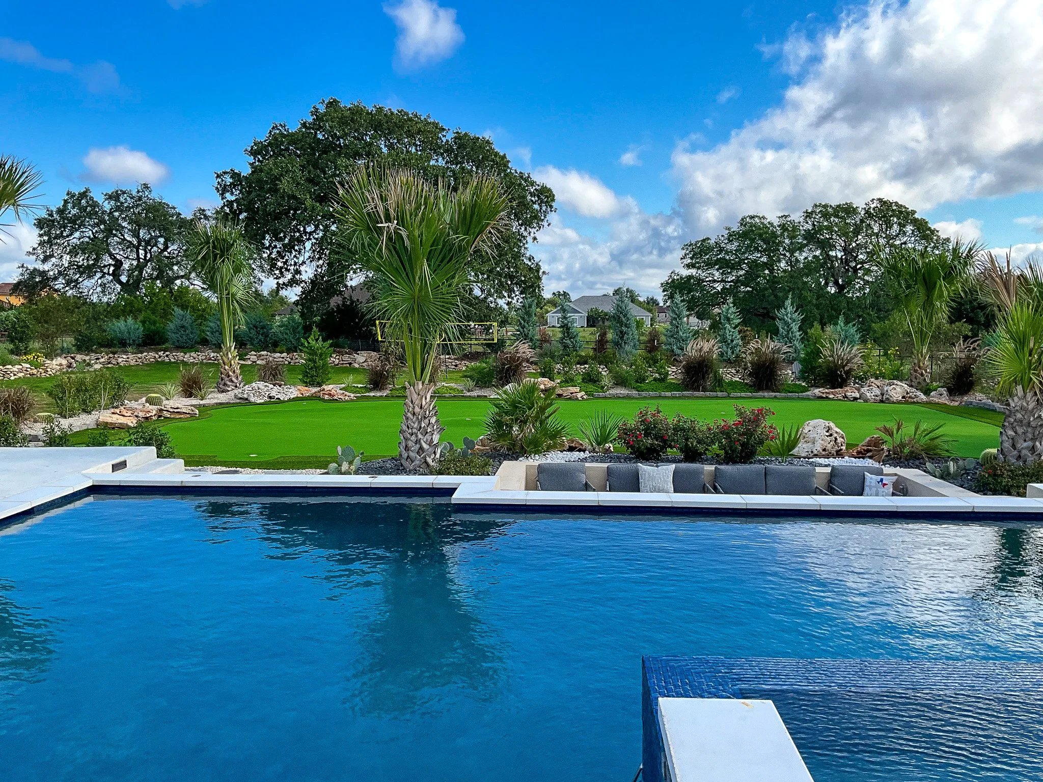 A backyard with a swimming pool in the foreground, surrounded by gray seating, with palm trees, green lawn, and landscaped garden with rocks and bushes under a partly cloudy sky.