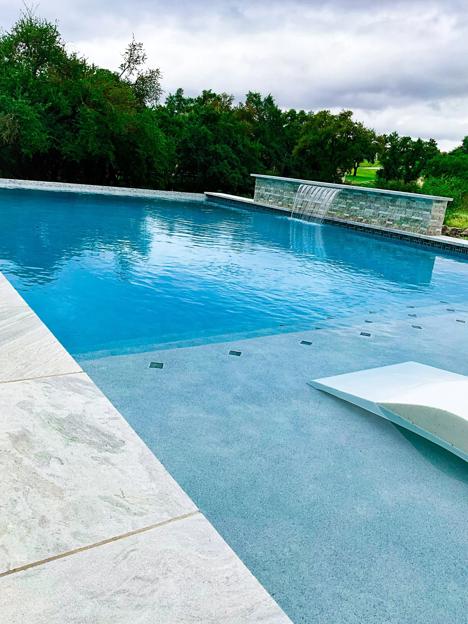 A clear blue swimming pool with a small waterfall feature in a lush green outdoor setting with trees and a cloudy sky in the background.