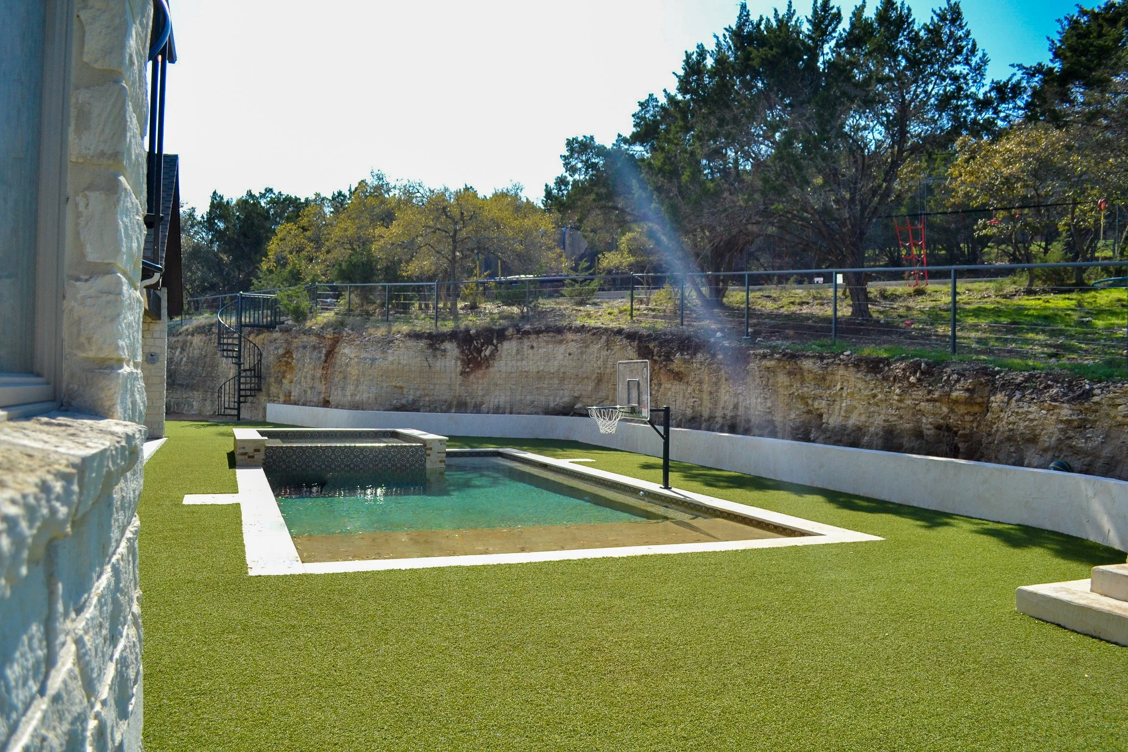 A backyard with a small pool, artificial grass, and a basketball hoop, with trees and fence in the background.