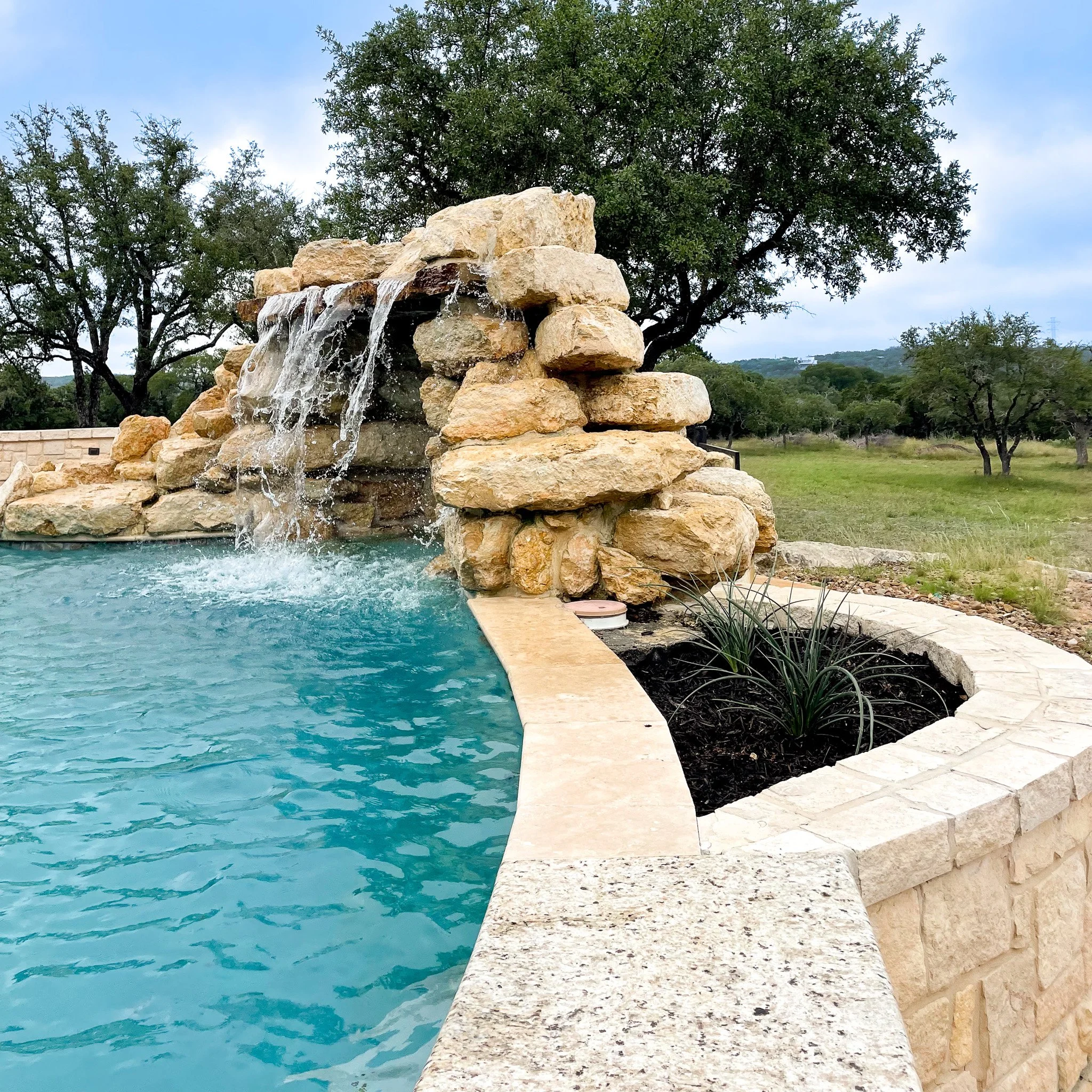 A swimming pool with a rock waterfall feature surrounded by a stone deck and landscaping, set in a grassy area with trees in the background.
