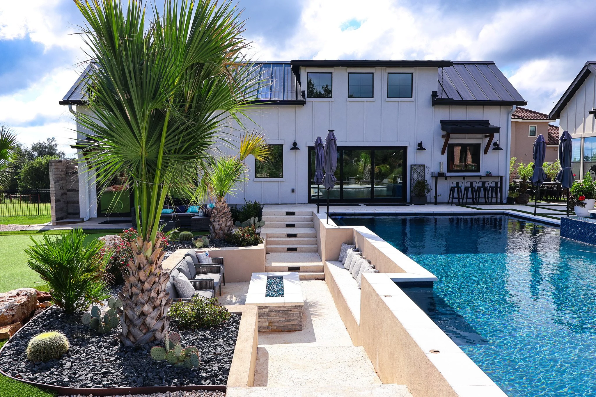Modern backyard with swimming pool, desert plants, and outdoor seating area in front of a white house with black accents.