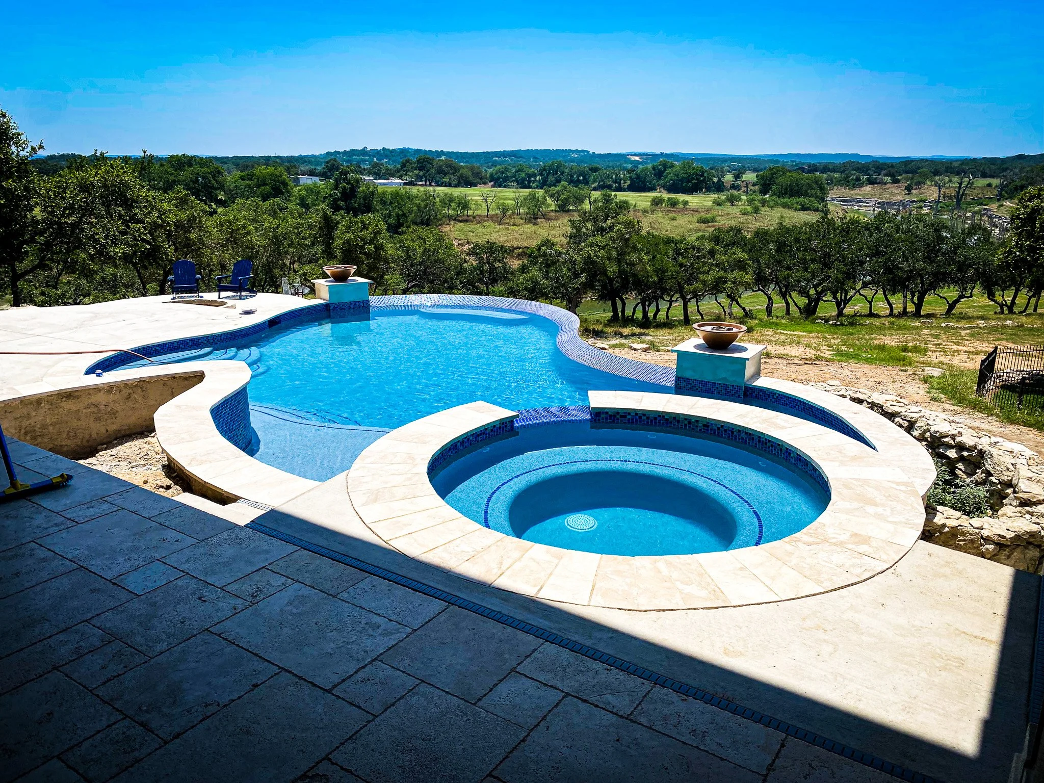 Swimming pool with hot tub in backyard overlooking trees and open countryside under a clear blue sky.