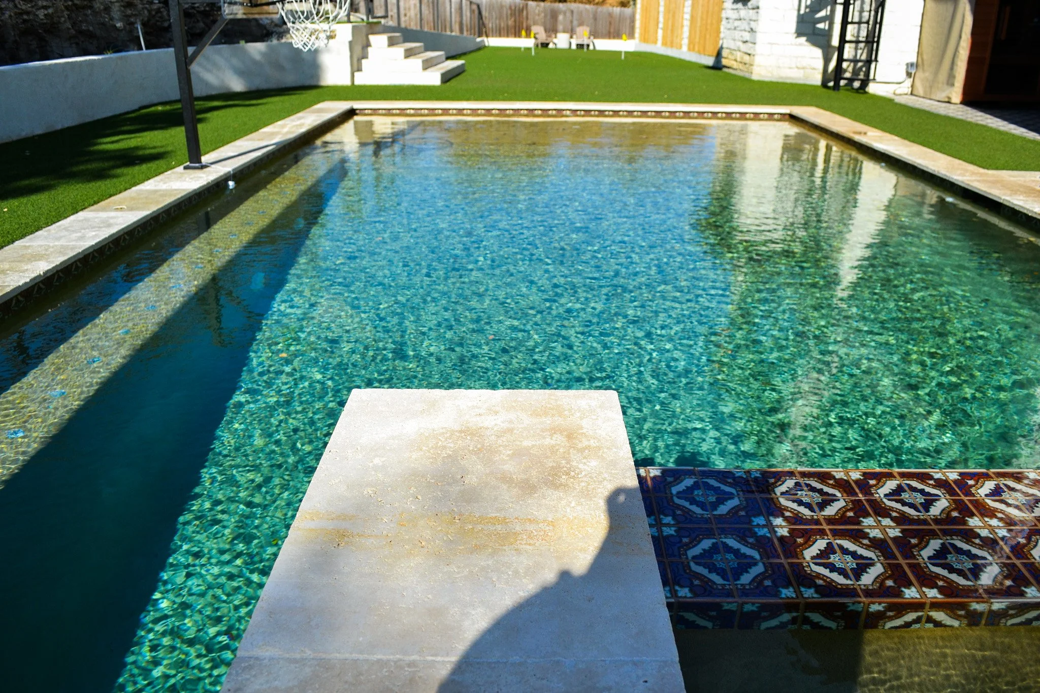 A backyard swimming pool with clear water, surrounded by green artificial grass, white concrete steps in the background, and decorative tiles at the edge of the pool, with some outdoor chairs and furniture on the patio.