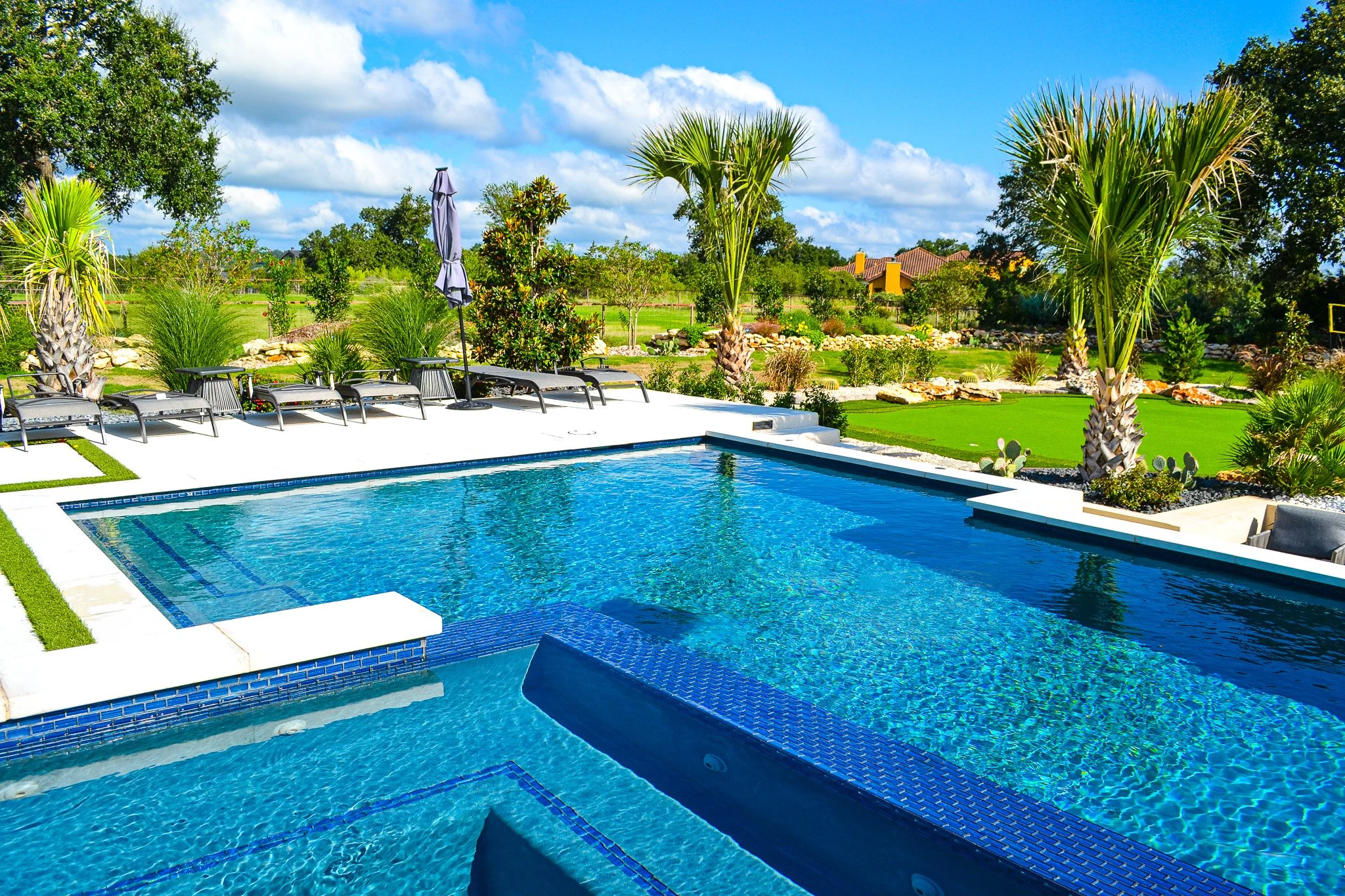 Luxury backyard swimming pool with lounge chairs, umbrellas, tropical trees, and lush greenery under a blue sky with white clouds.