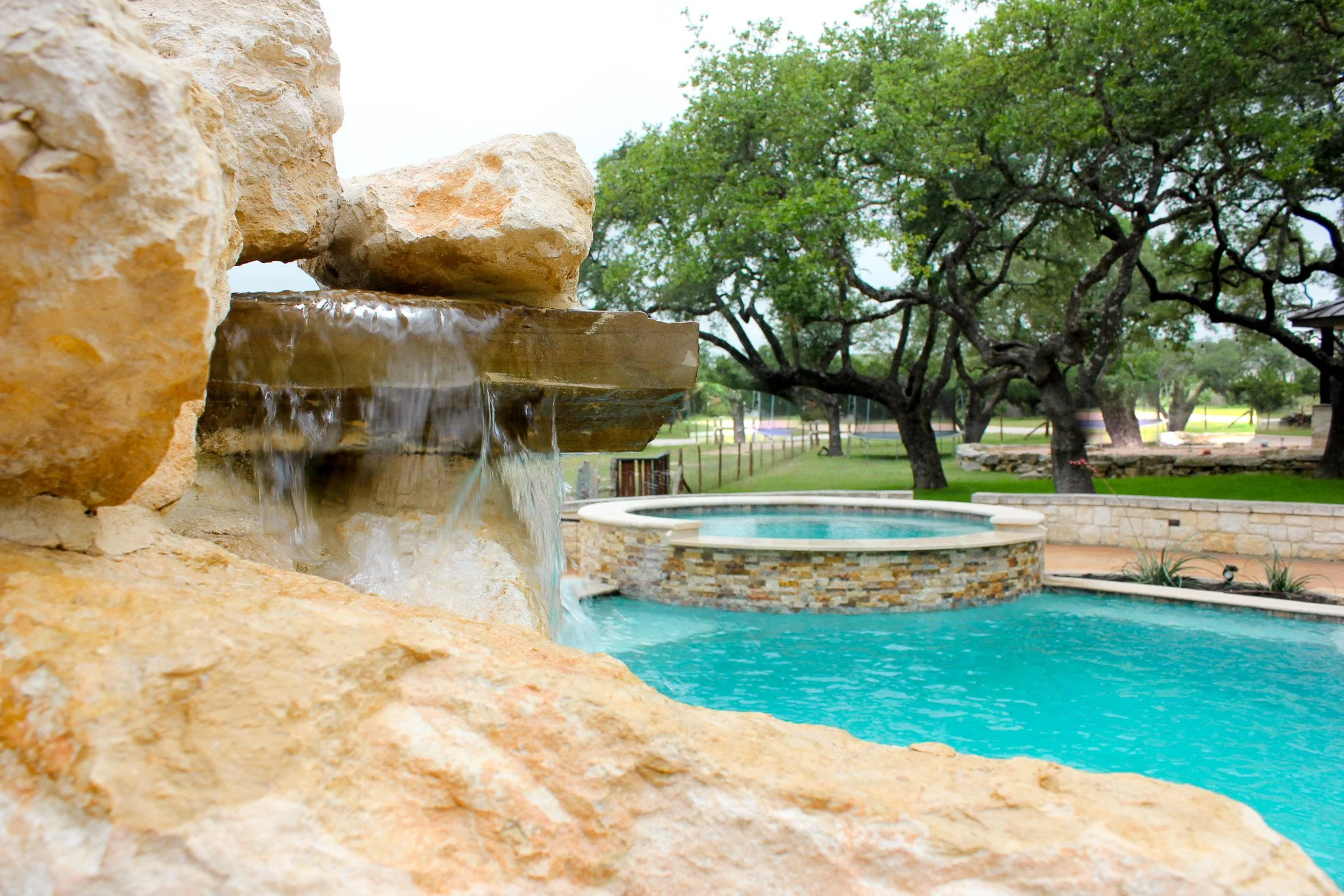 View of a backyard with a swimming pool and a water feature made of rocks, a stone fire pit, mature trees, and a grassy landscape.