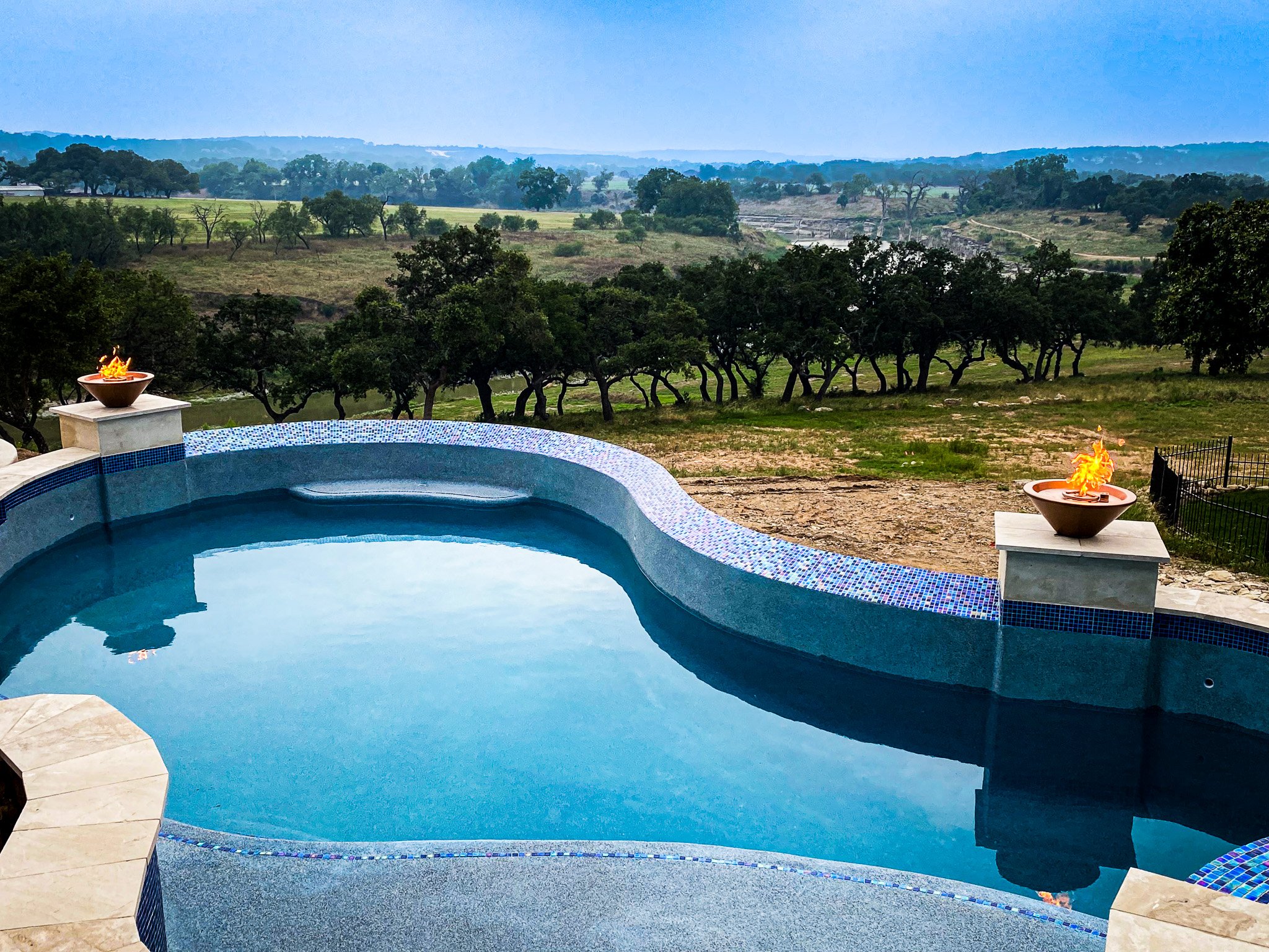 Infinity pool with two fire bowls overlooking a rural landscape with rolling hills, trees, and a clear blue sky.