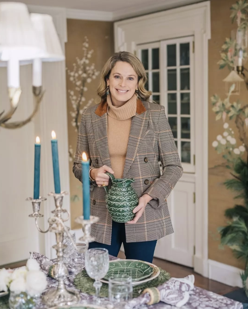 A smiling woman holding a green ceramic pitcher in a decorated dining room with candles, tableware, and holiday decorations.