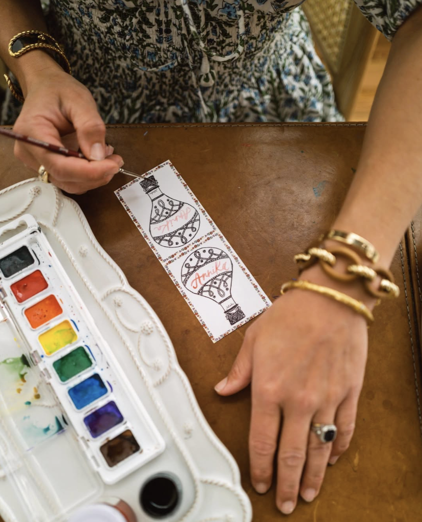 Person painting a decorative ornament that says 'Thank You' on a strip of paper, with watercolor paints and brushes on a white tray, wearing gold jewelry, sitting at a wooden table.