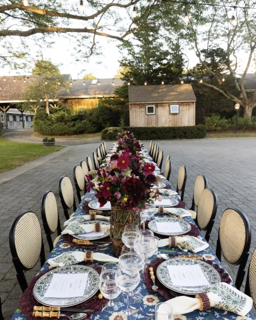 A long outdoor dining table decorated with floral centerpieces, wine glasses, plates, and napkins, set in a garden with string lights overhead and a small wooden building in the background.
