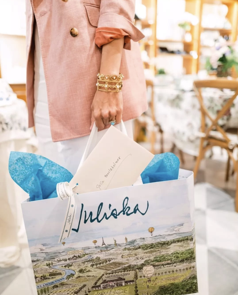 A person holding a shopping bag with the name Juliska, inside a well-lit store. The person is wearing a pink blazer and several bracelets.