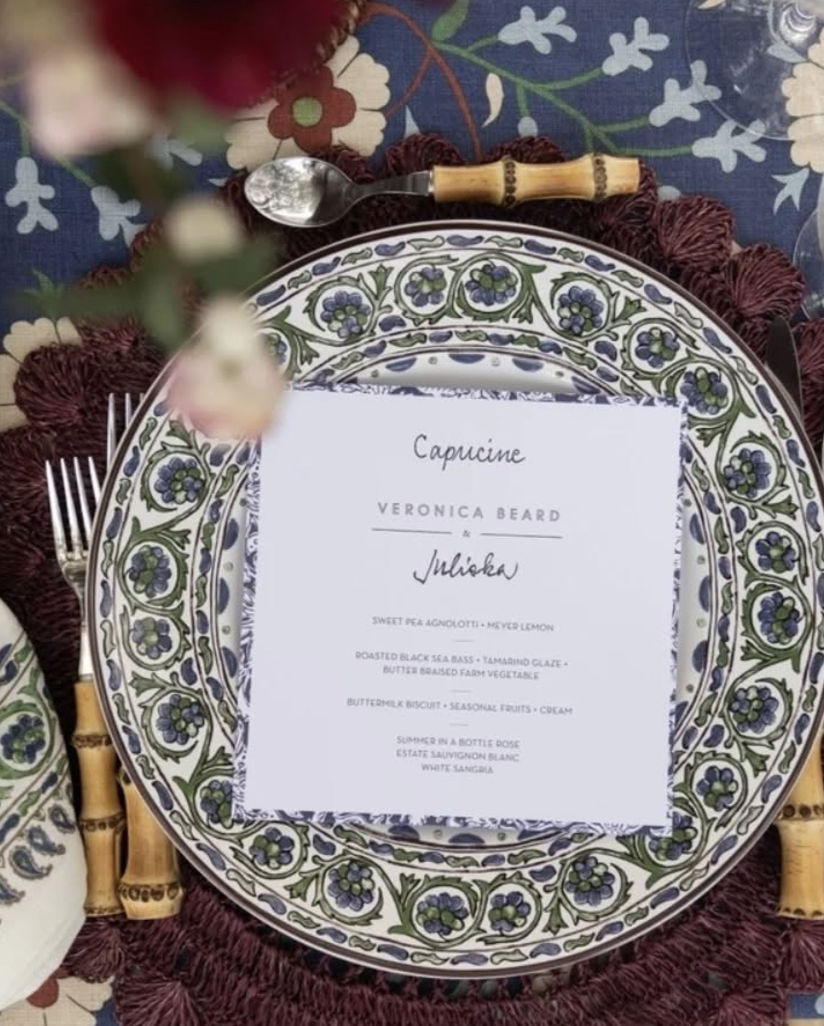 Elegant table setting with a decorated plate, a printed menu, a spoon with a bamboo handle, and floral tablecloth in the background.