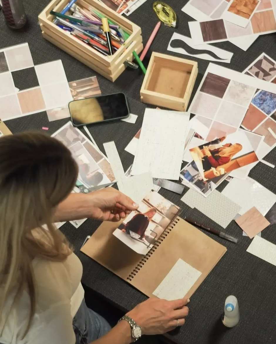 A woman working on a scrapbook or photo album on a black surface, surrounded by printed photos, colorful pens, a yellow tape measure, and various paper sheets and sample swatches.