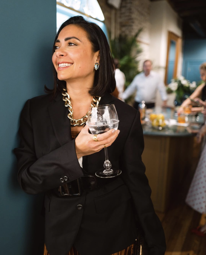 Woman smiling in a black suit, holding a glass of water, at a social gathering or party.