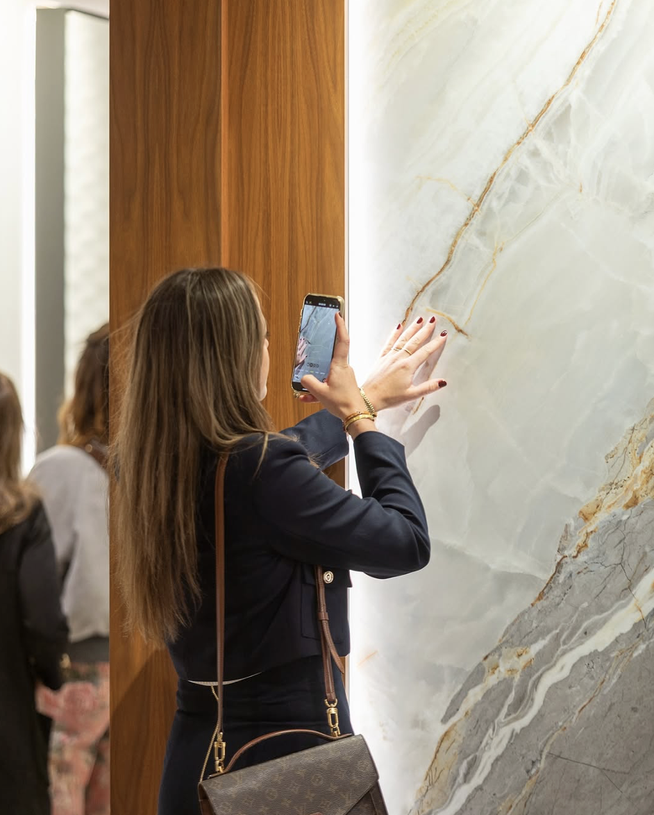 A woman with long brown hair in a black blazer taking a photo with her smartphone of a marble wall in a modern interior.