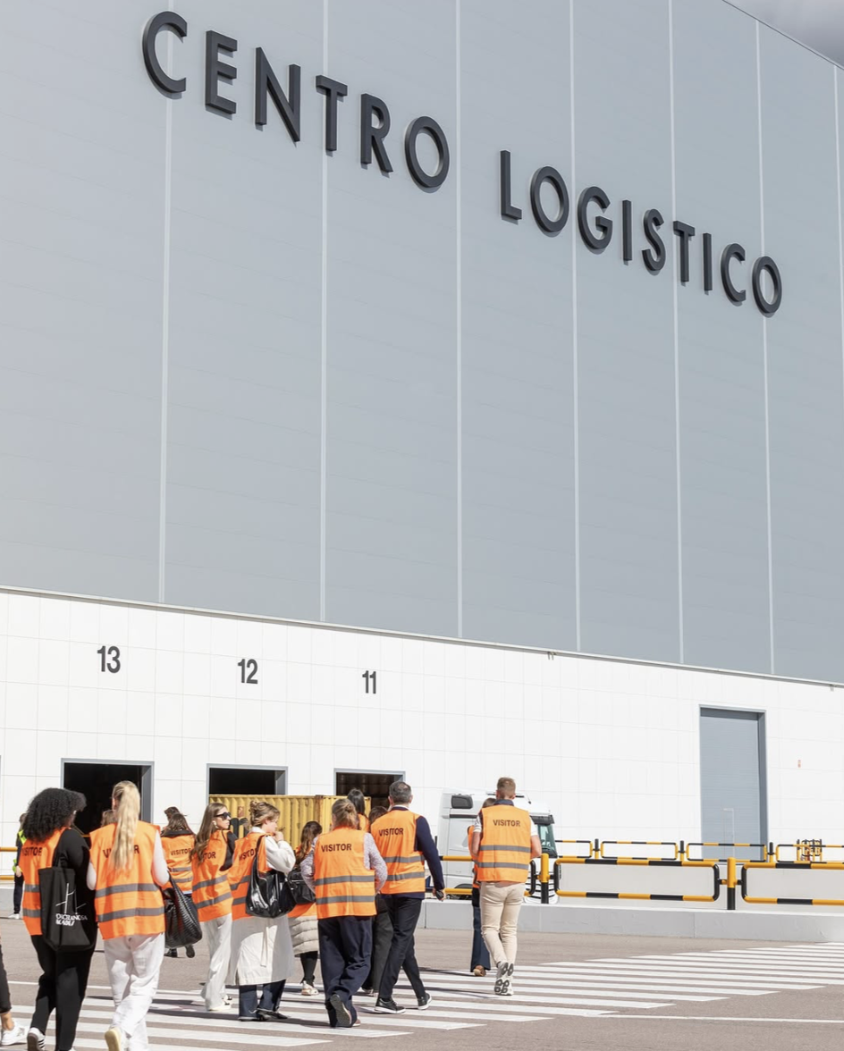 Group of visitors wearing orange vests labeled 'visitor' entering a large logistics center with the sign 'Centro Logistico' on the building.
