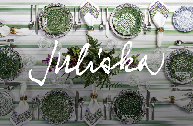 A festive table setting with green and white patterned plates, silverware, and white napkins with green and purple napkin rings, arranged above a striped tablecloth with the word 'Jubilosa' written across the center.