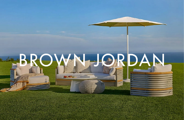 Outdoor seating area with cushioned chairs and a table on green grass, under a large white patio umbrella, overlooking the ocean on a clear day.