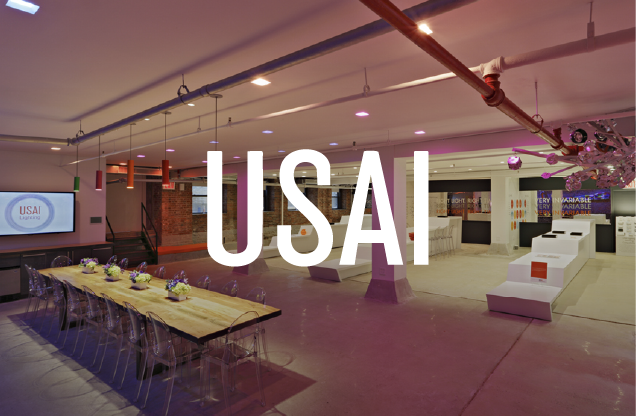 Event space with a long table set with flowers, chairs, and a large screen displaying 'USA' and 'USAI' logo, with a decorated lounge area and pink balloons in the background.