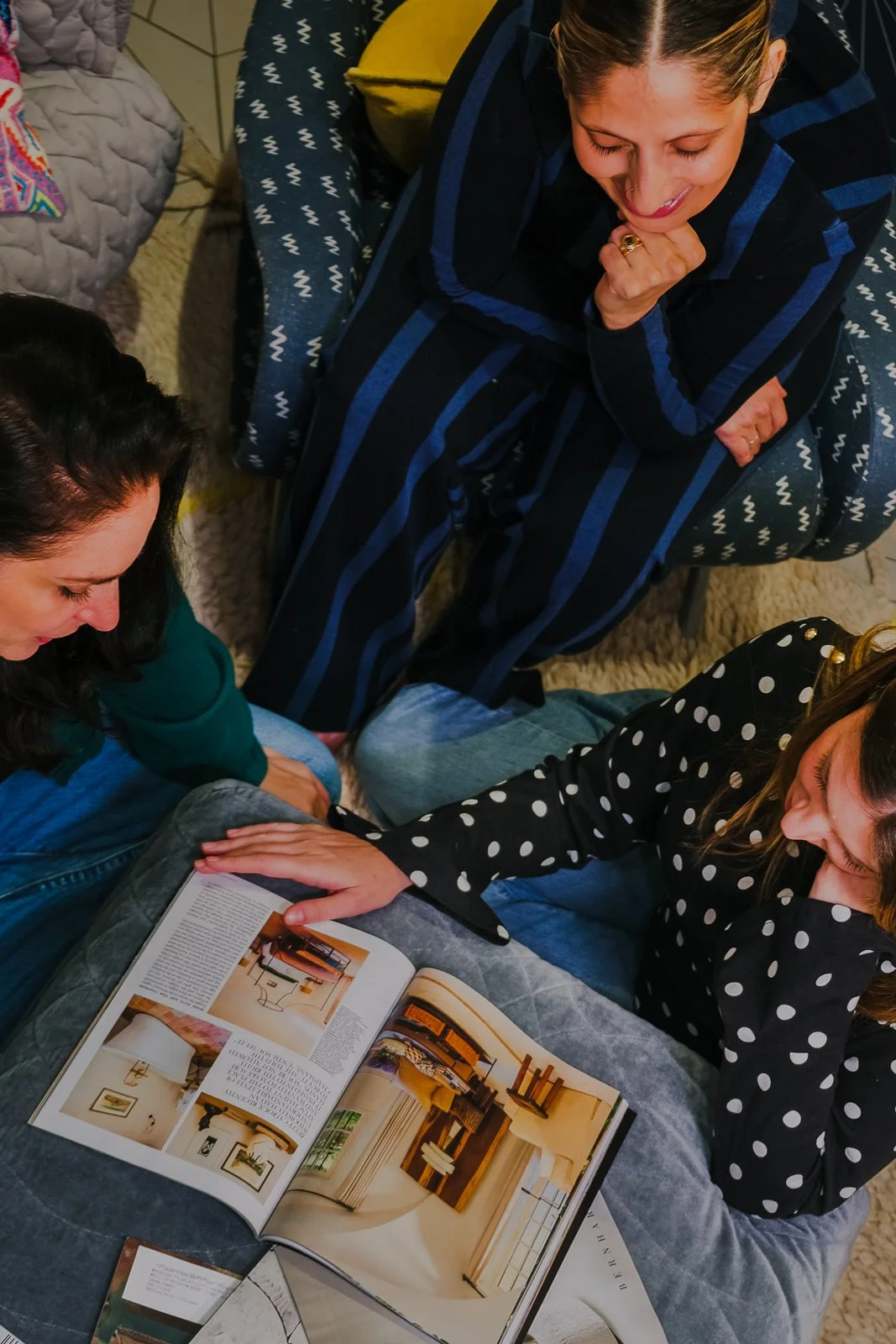 Three women sitting on the floor and looking at an open design magazine. One woman is pointing at an interior page, while the other two women are engaged and watching.
