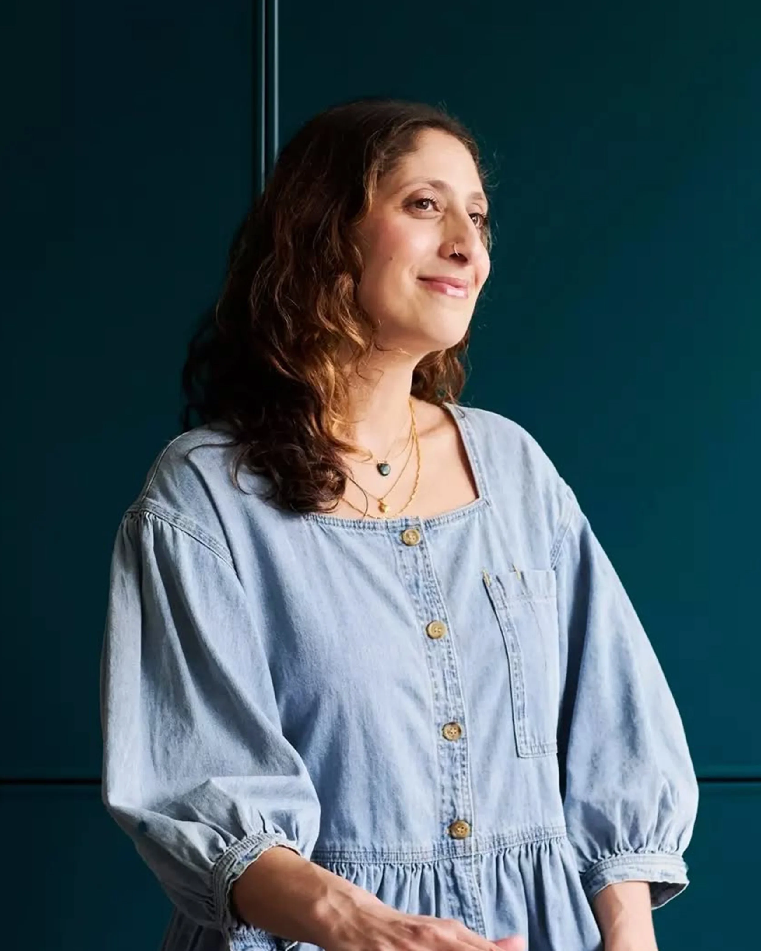 A woman with curly brown hair wearing a light blue denim dress with buttons, layered necklaces, and a nose piercing, standing against a dark teal background.