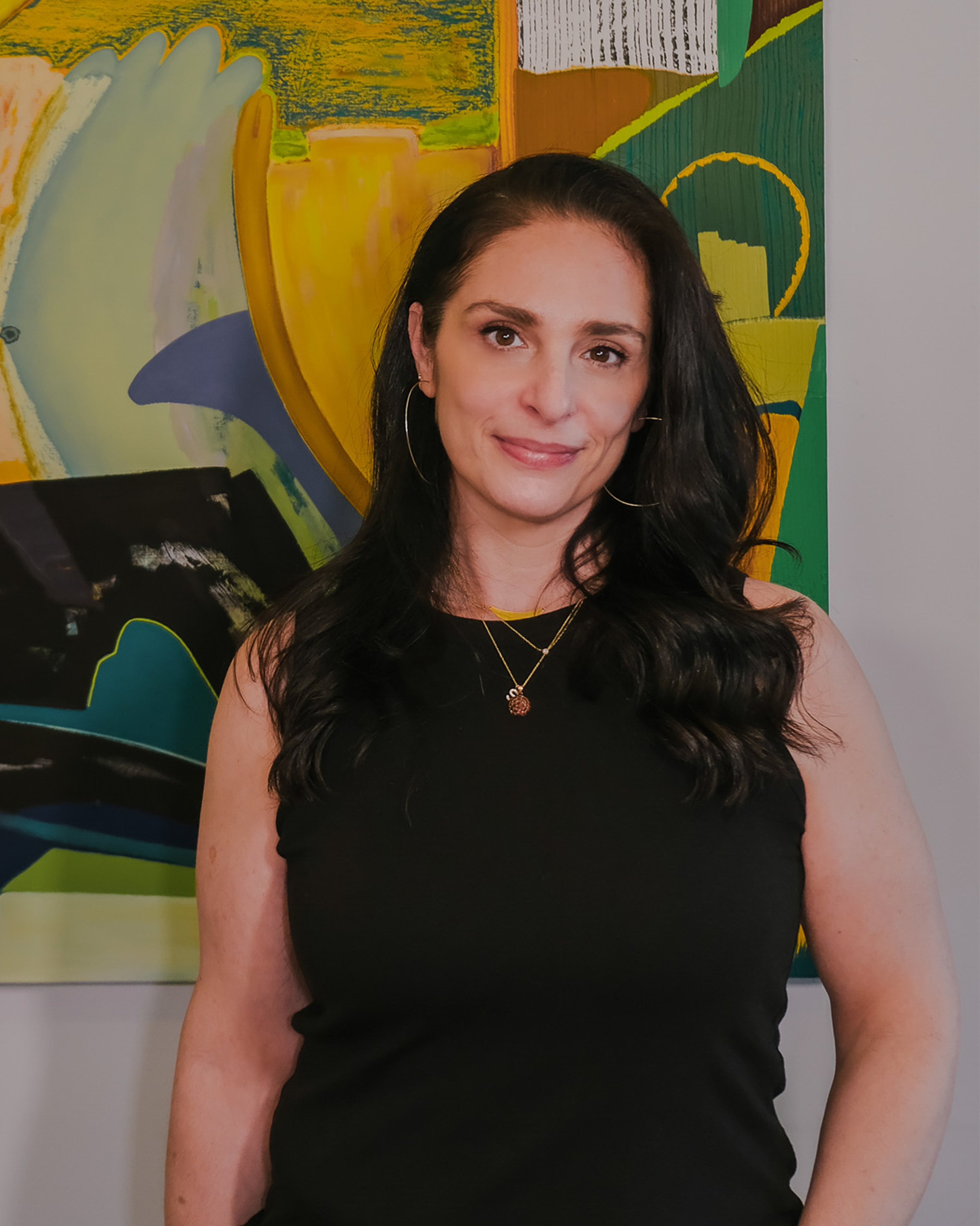 A woman with long dark hair, wearing hoop earrings and layered necklaces, smiles at the camera. She is dressed in a black sleeveless top and stands in front of a colorful abstract painting.