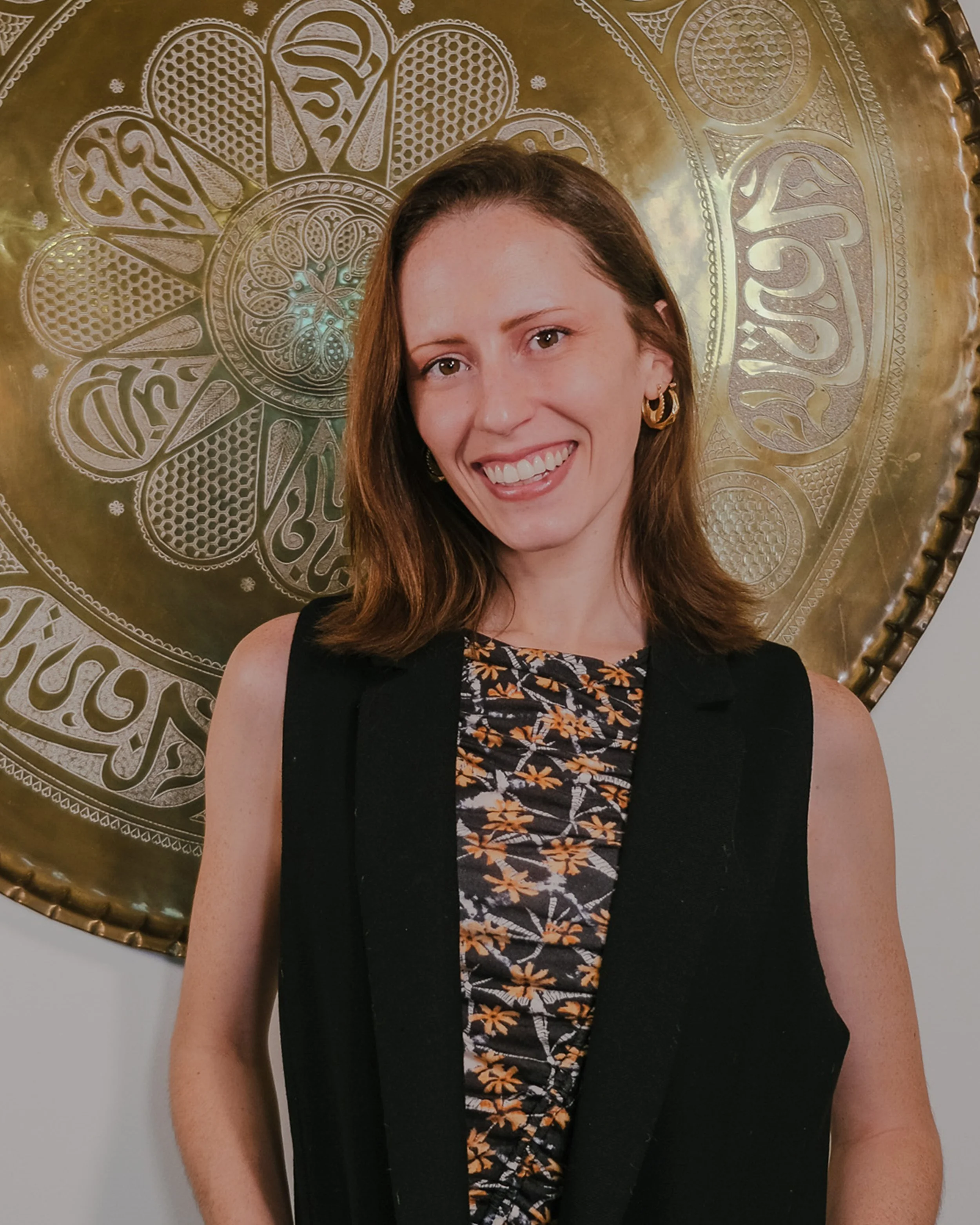 A woman with shoulder-length brown hair, wearing a black vest over a floral dress, stands in front of a large decorative brass plate wall art, smiling at the camera.