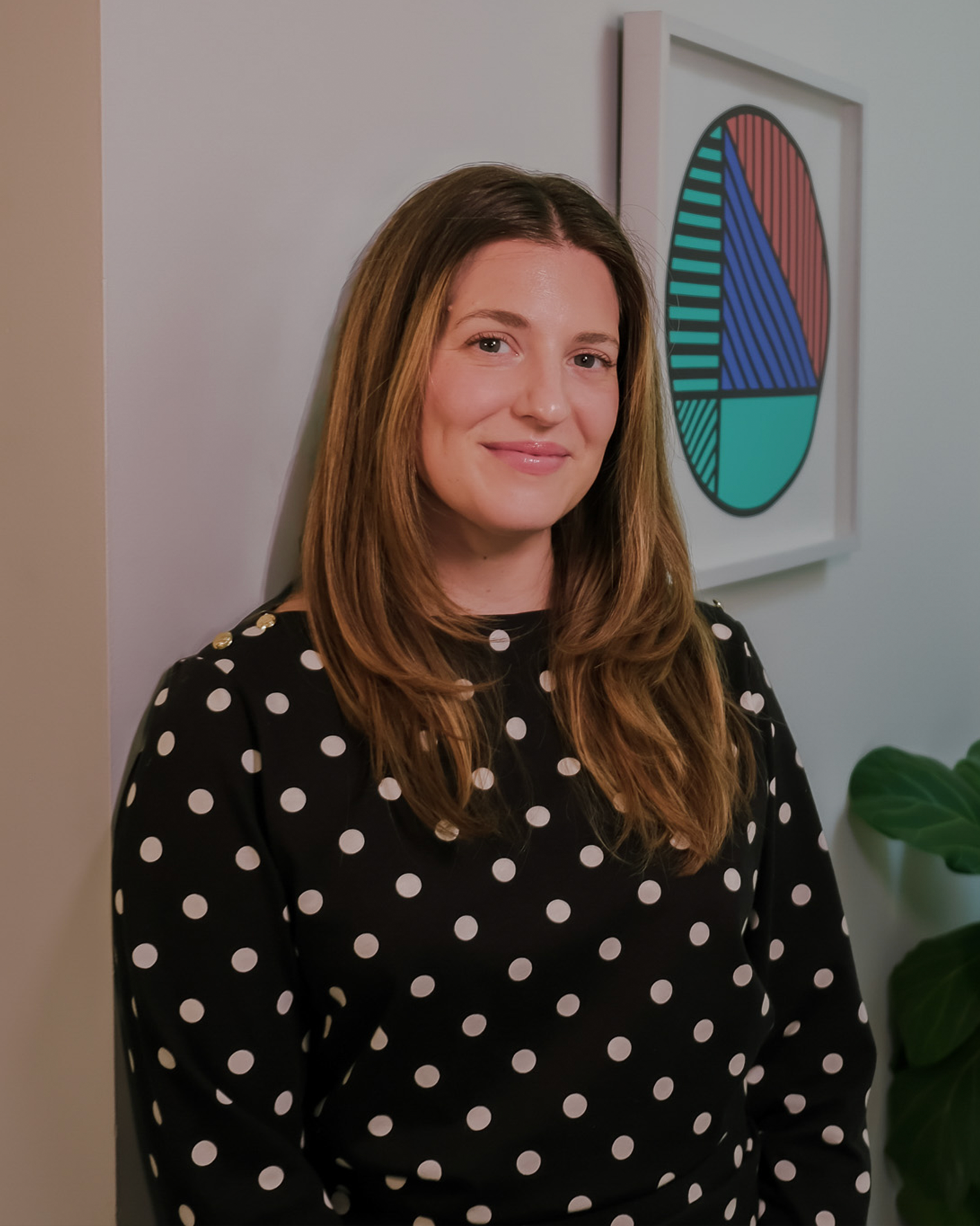 A young woman with long, wavy brown hair smiling, wearing a black dress with white polka dots, standing indoors in front of a white wall with a framed colorful geometric art piece, and a large green plant.