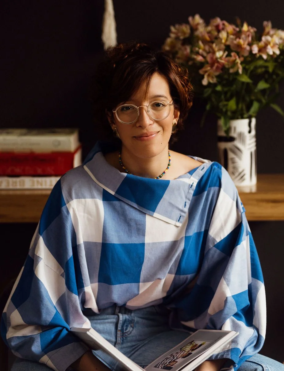 Woman wearing glasses and a blue checkered shirt sitting indoors with a book, flowers in a vase behind her, and a dark background.