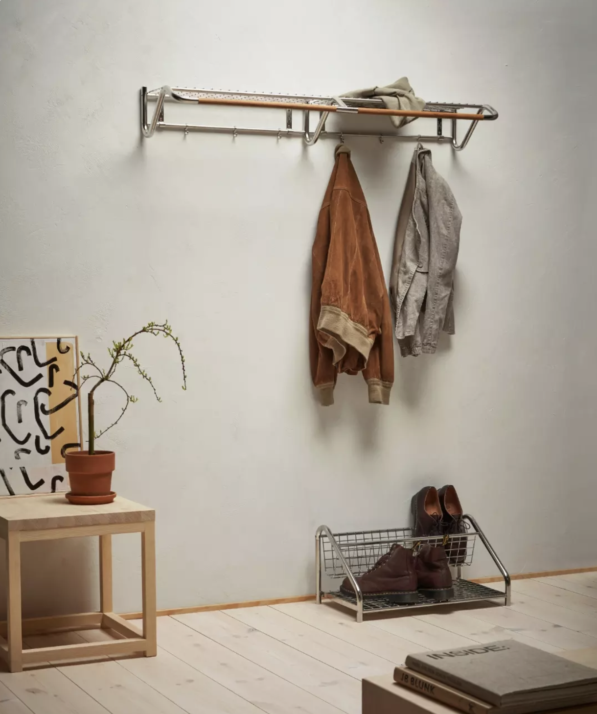 Entryway with a coat rack holding a brown jacket and a gray shirt, a shoe rack with brown boots, a small wooden table with a potted plant, and a framed abstract artwork leaning against the wall.