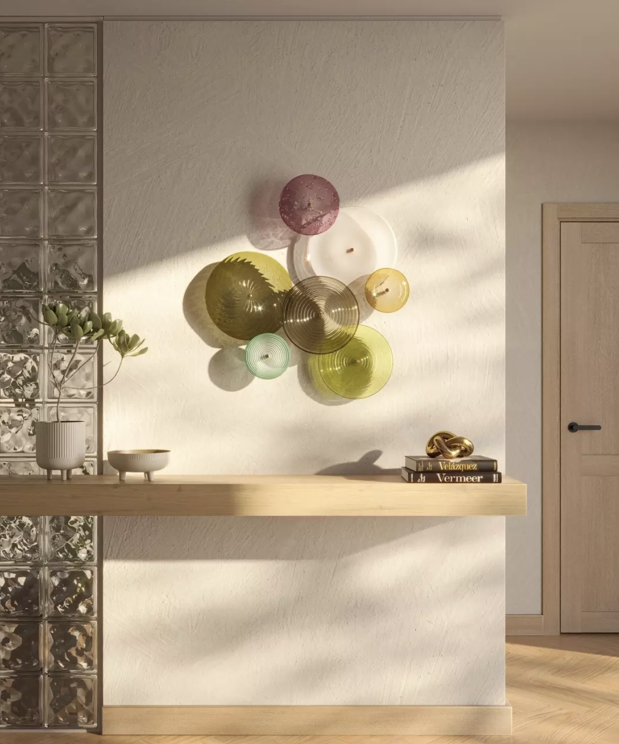 Decorative wall art composed of colored glass or plastic discs on a white wall, illuminated by sunlight. A wooden shelf below holds a potted plant, two bowls, a stack of books, and a decorative gold sculpture. Part of a glass block wall is visible on the left, and a wooden door is on the right.