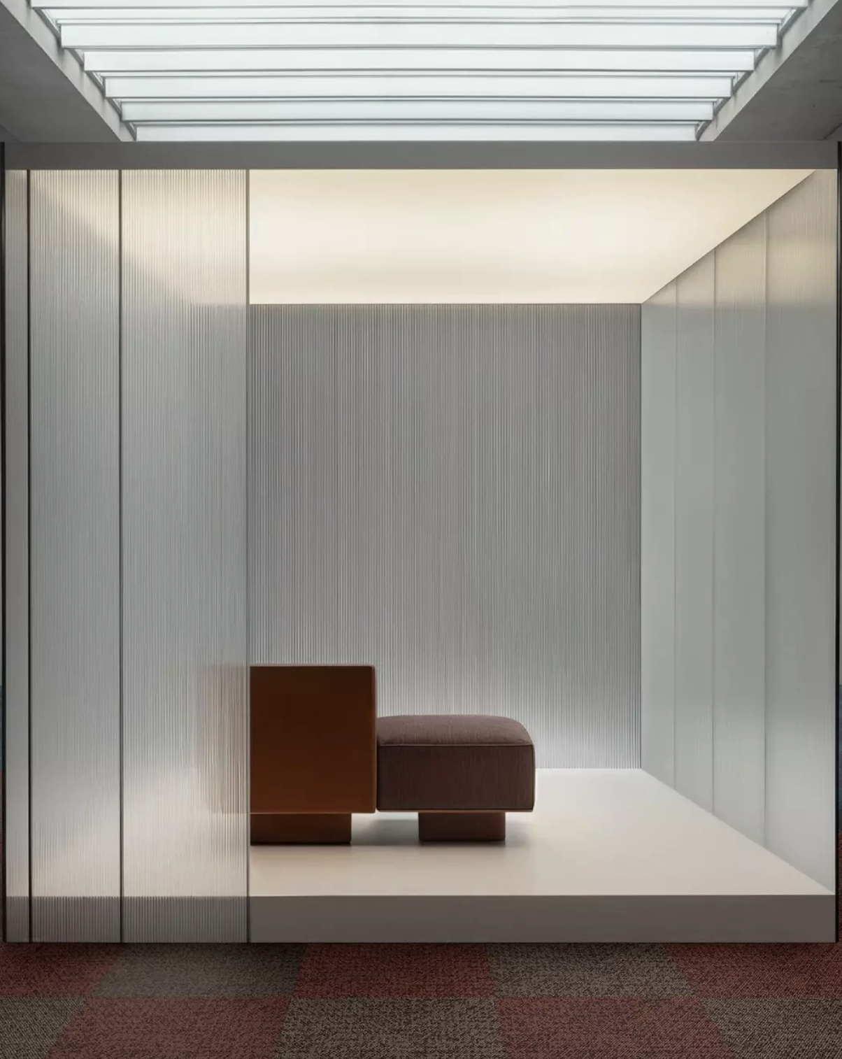 Modern interior with a brown and maroon lounge chair within a minimalist, well-lit room featuring vertical textured wall panels and a ceiling with frosted glass or translucent panels.
