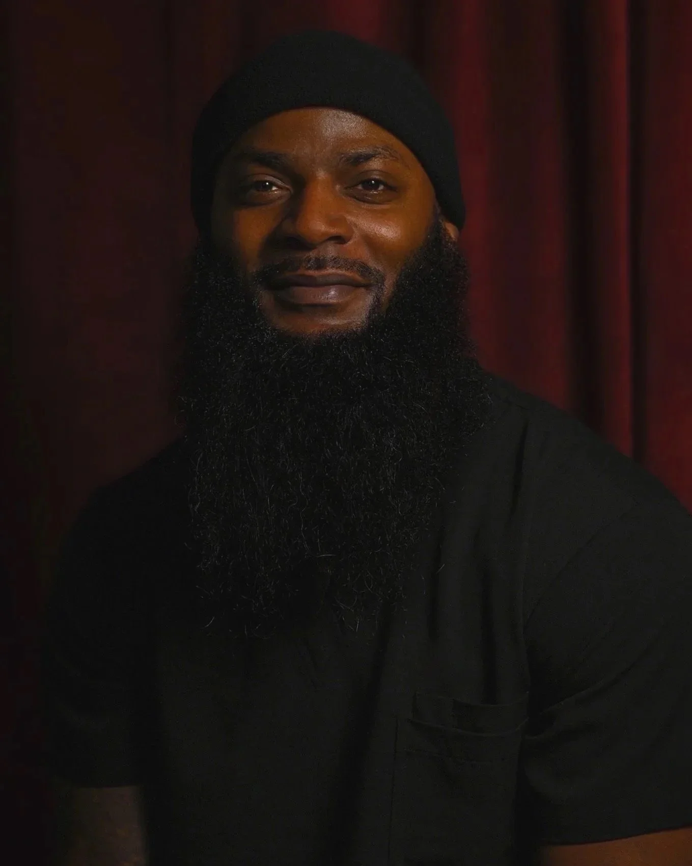 A man with a full beard wearing a black beanie and black shirt, smiling slightly, standing in front of a dark red curtain.