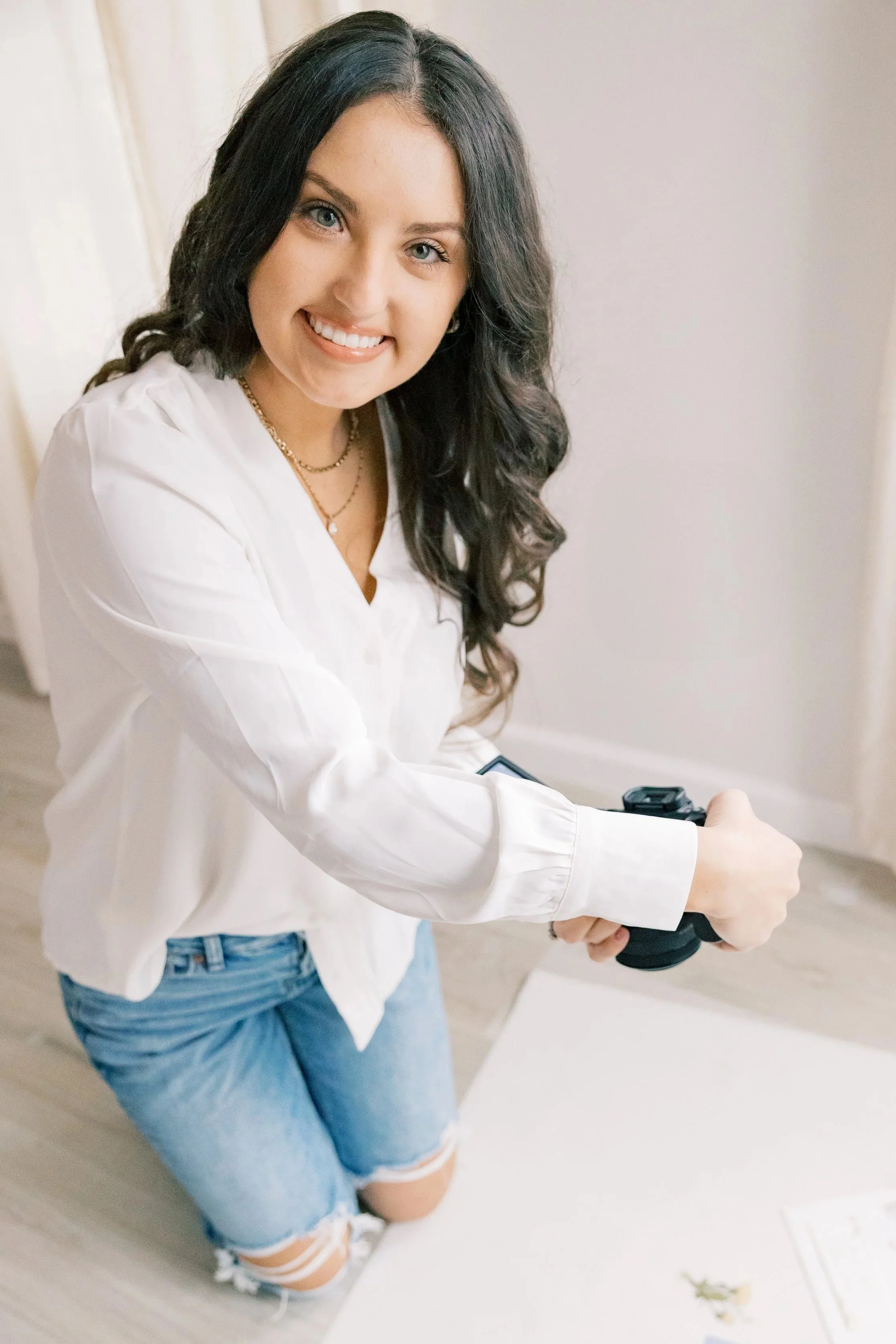 Kenzie, of Kenzie Riley Photography from Kentucky with long dark hair, wearing a white shirt and ripped jeans, kneeling on the floor and smiling while holding a camera.