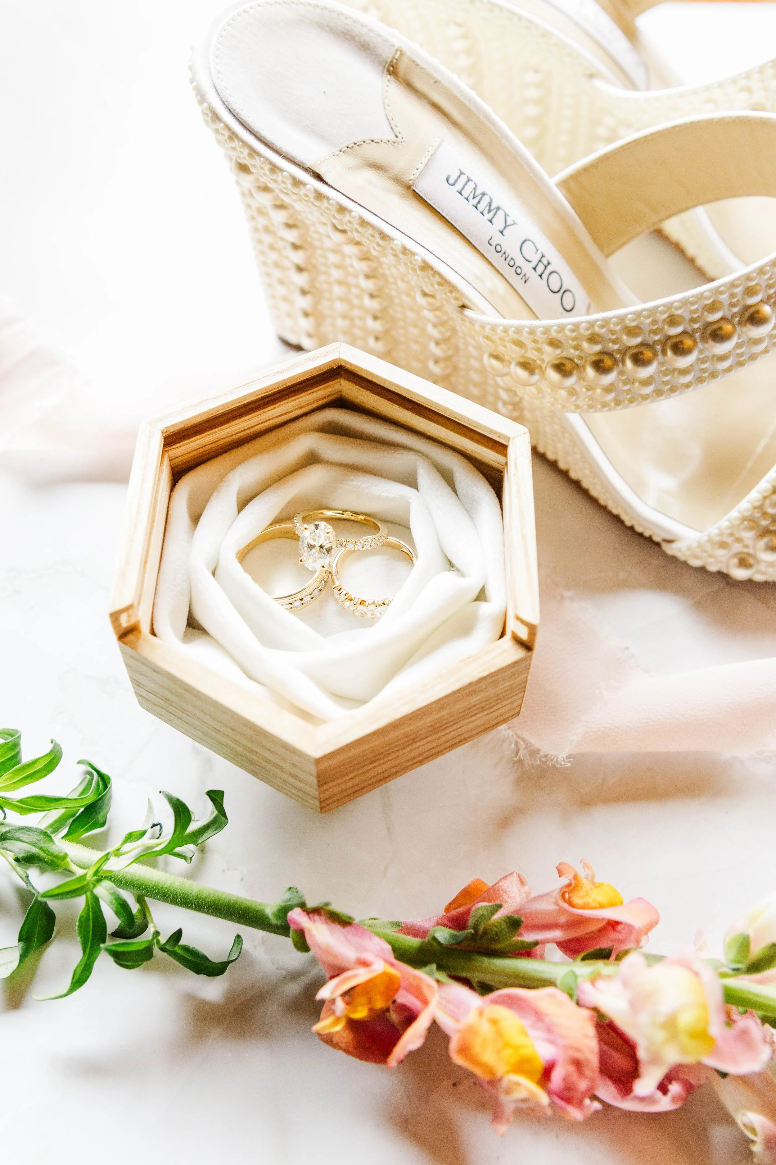 A pair of Jared engagement rings in a wooden ring box with white velvet interior, a pair of white high-heeled shoes by Jimmy Choo with pearl and bead embellishments, and pink and orange flowers. Red Gate Farm and Vineyard, Quincy, KY.