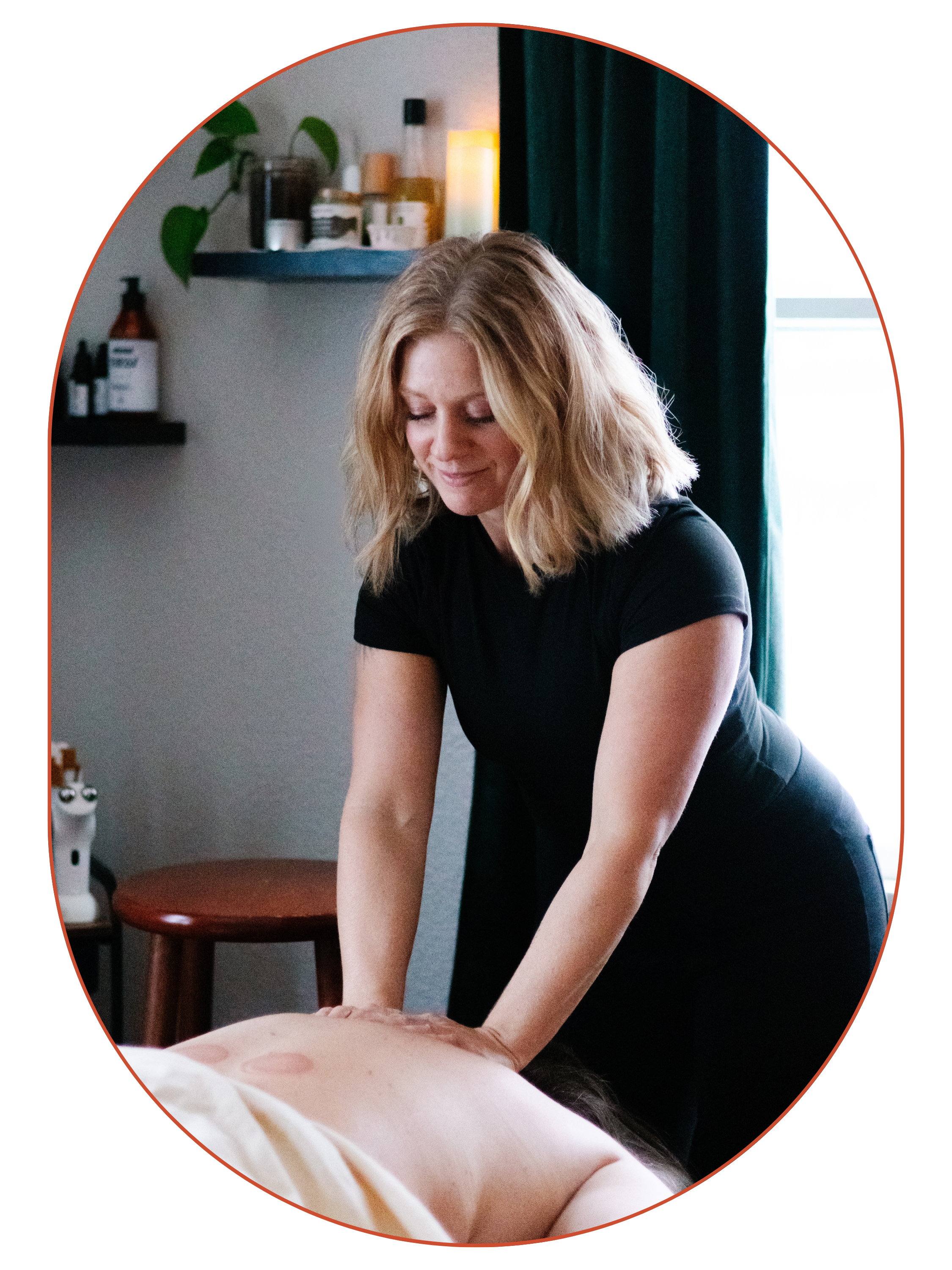 A woman giving a massage to a person lying face down on a massage table in a room with shelves containing bottles and a plant.