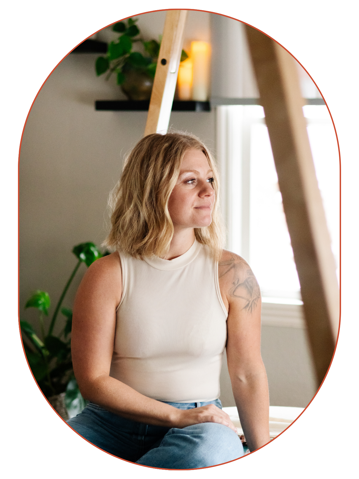 A woman with blonde, wavy shoulder-length hair sits on a surface near a window, looking thoughtfully to the side. She wears a sleeveless cream-colored top and light blue jeans, with a tattoo on her upper left arm. The background features a plant, a dark shelf with candles, and a window allowing natural light to fill the room.