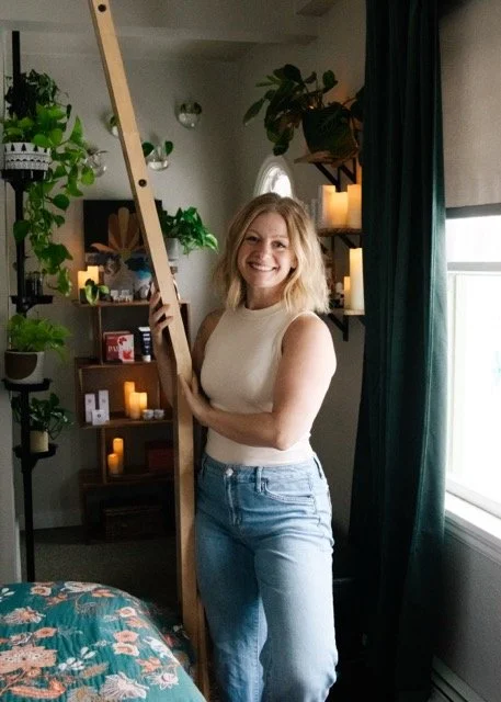 Woman with blonde hair smiling and holding a ladder, standing in a room with green plants, candles, and books on shelves, near a window with dark curtains.