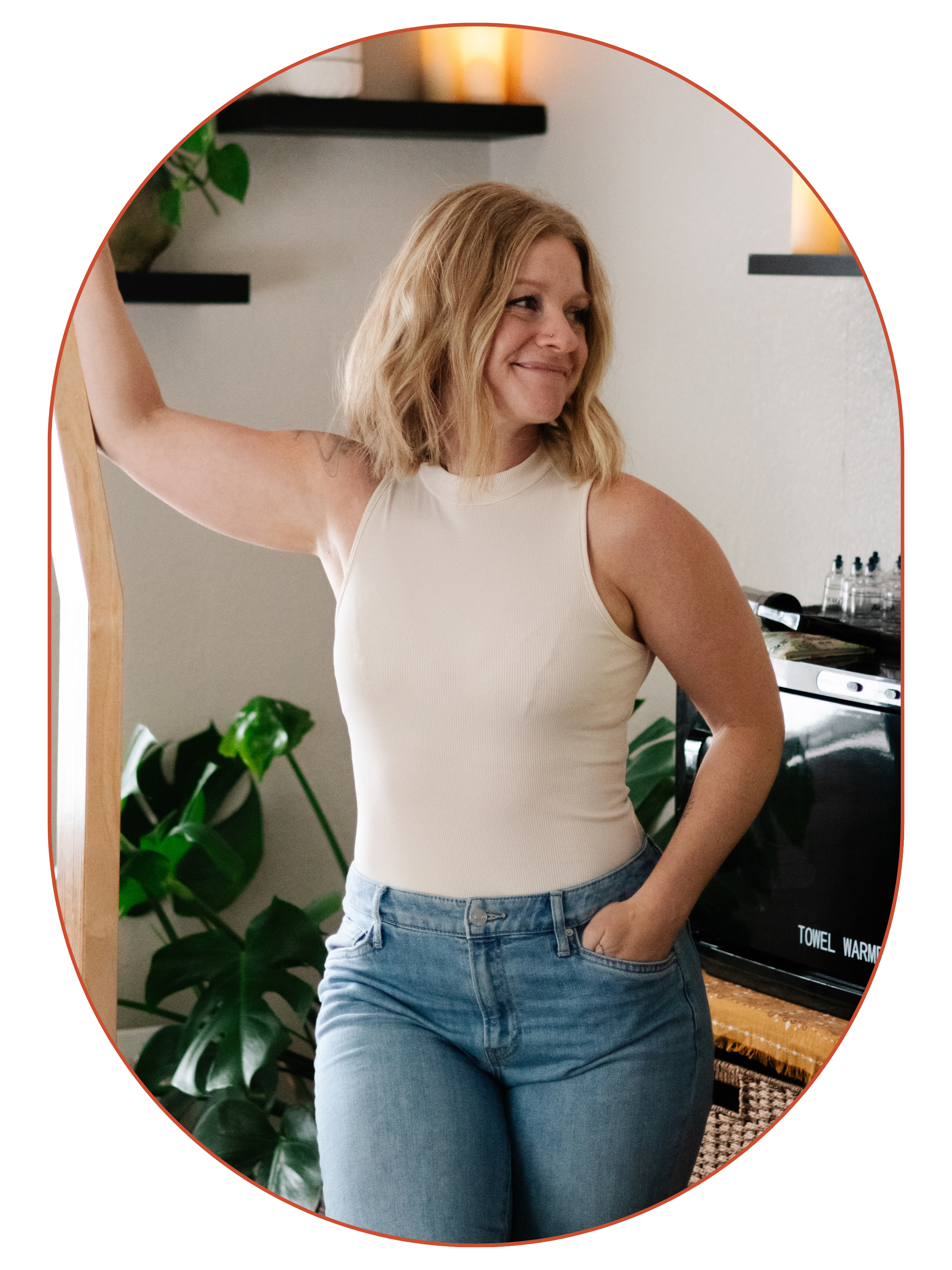 A woman with shoulder-length blond hair smiling and standing in a kitchen, wearing a sleeveless cream top and jeans, with her left hand in her pocket and her right arm resting on a wooden frame.