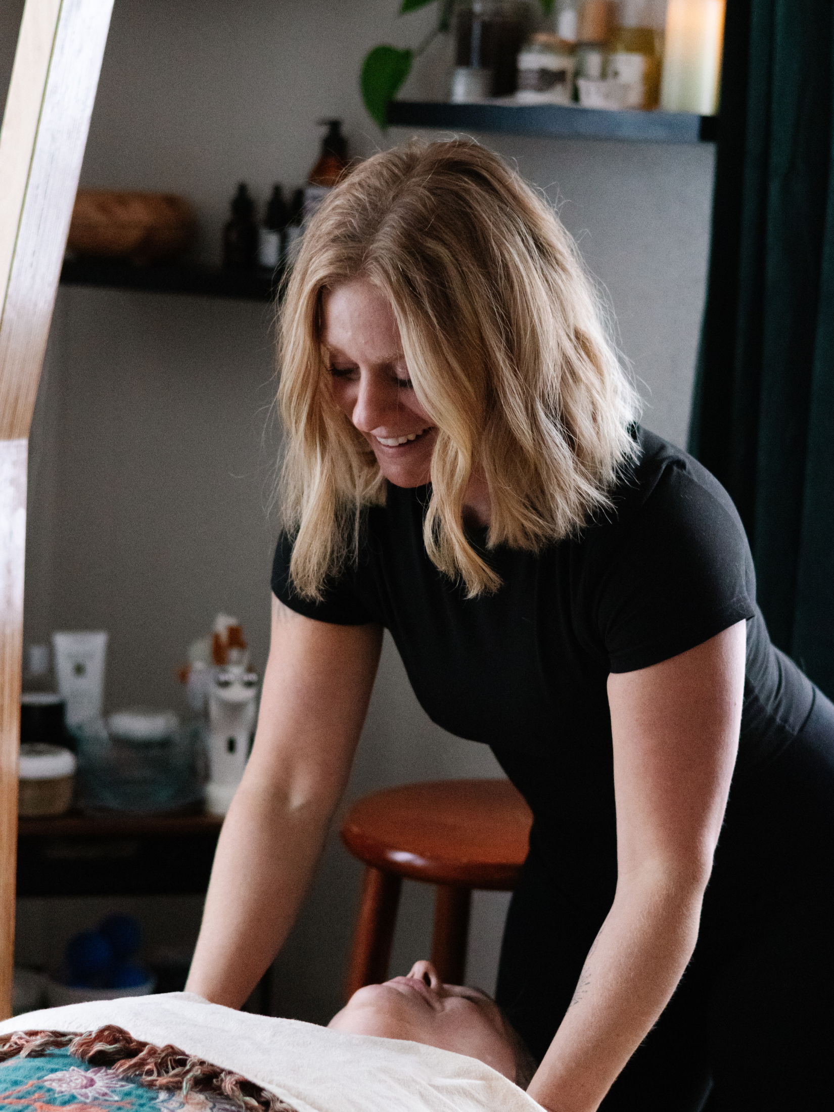 A woman with shoulder-length blonde hair, wearing a black shirt, is performing a massage on a person lying on a massage table in a wellness room.