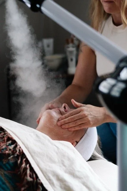 A woman receiving a facial treatment from a skincare professional in a spa or clinic setting, with steam or vapor in the air.