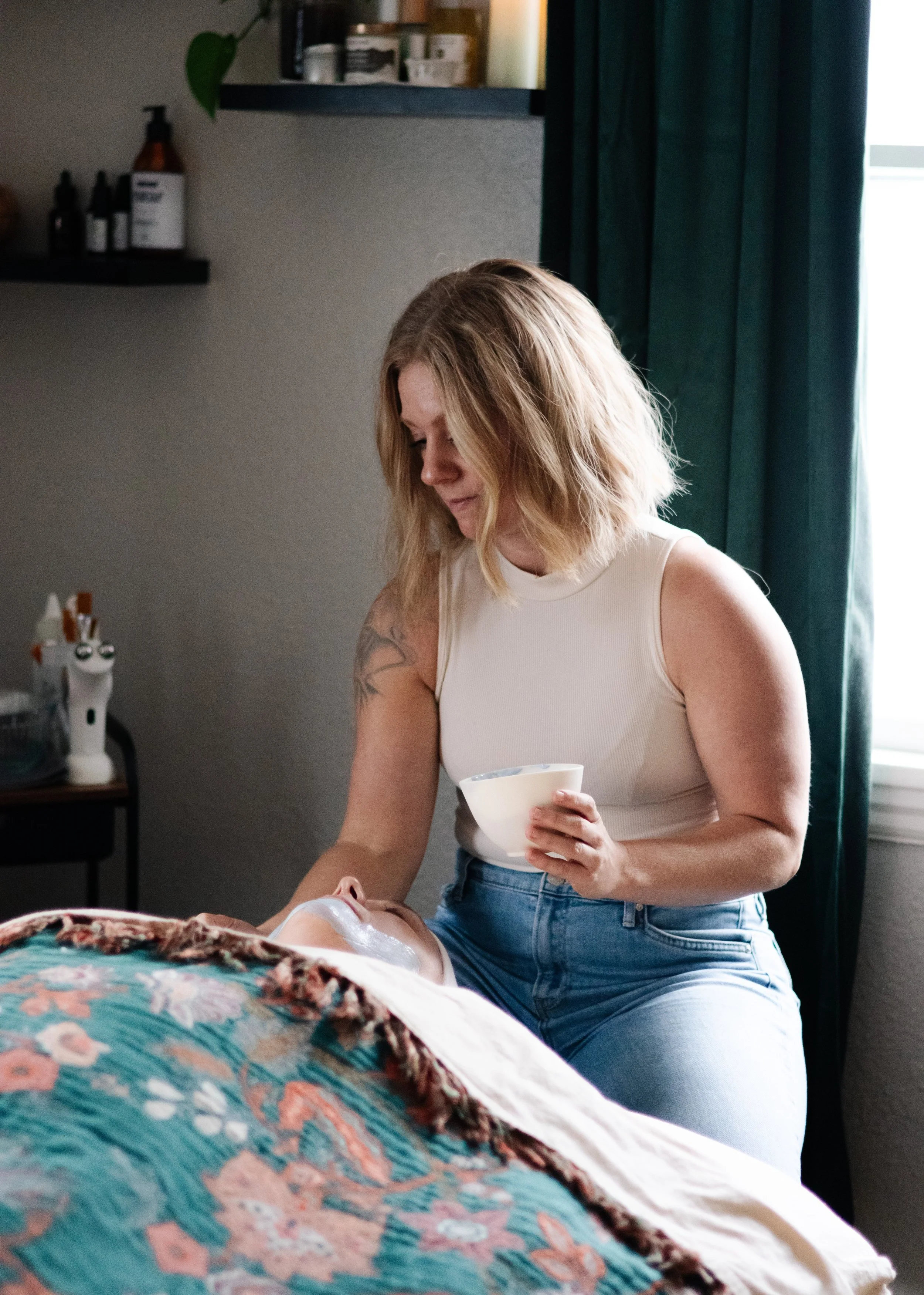 A woman with blonde hair giving a facial to a person lying down with a floral blanket, holding a bowl of facial products.
