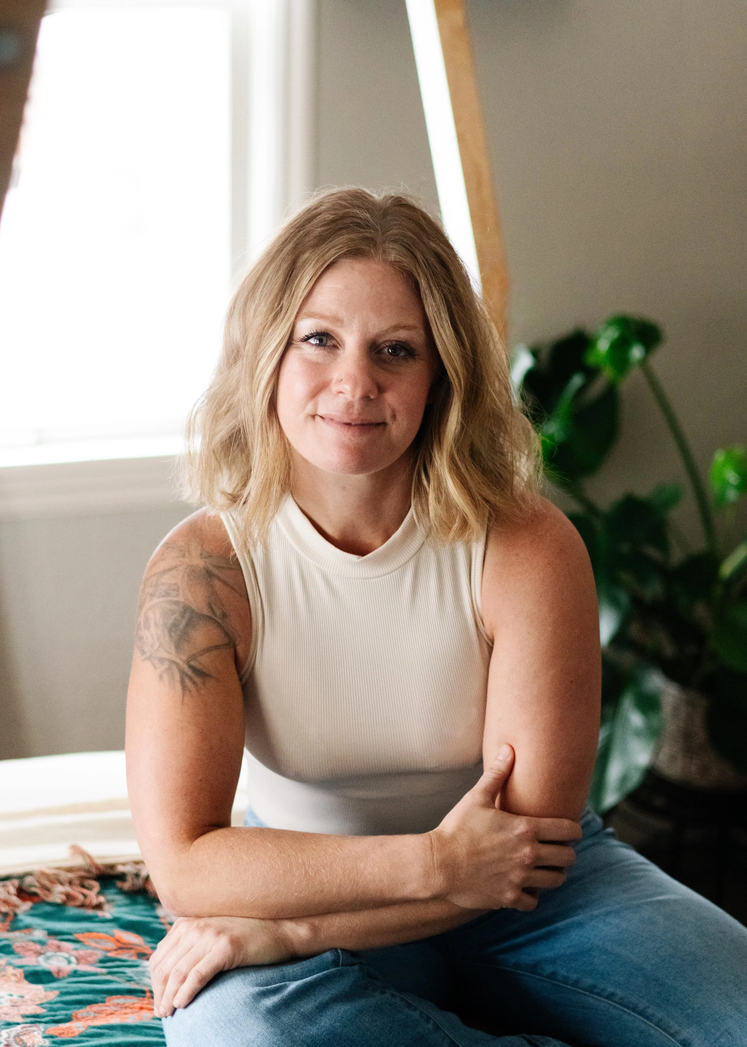 A woman with shoulder-length blonde hair, wearing a white sleeveless top and blue jeans, sitting at an angle on a bed with a colorful quilt, in a room with natural light from a window and a green potted plant in the background.