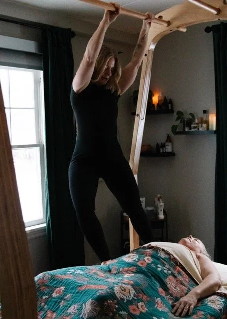 A woman in black workout clothes hanging upside down from a wooden ceiling support in a room with dark curtains and shelves, with a person lying on a bed with a colorful floral blanket.
