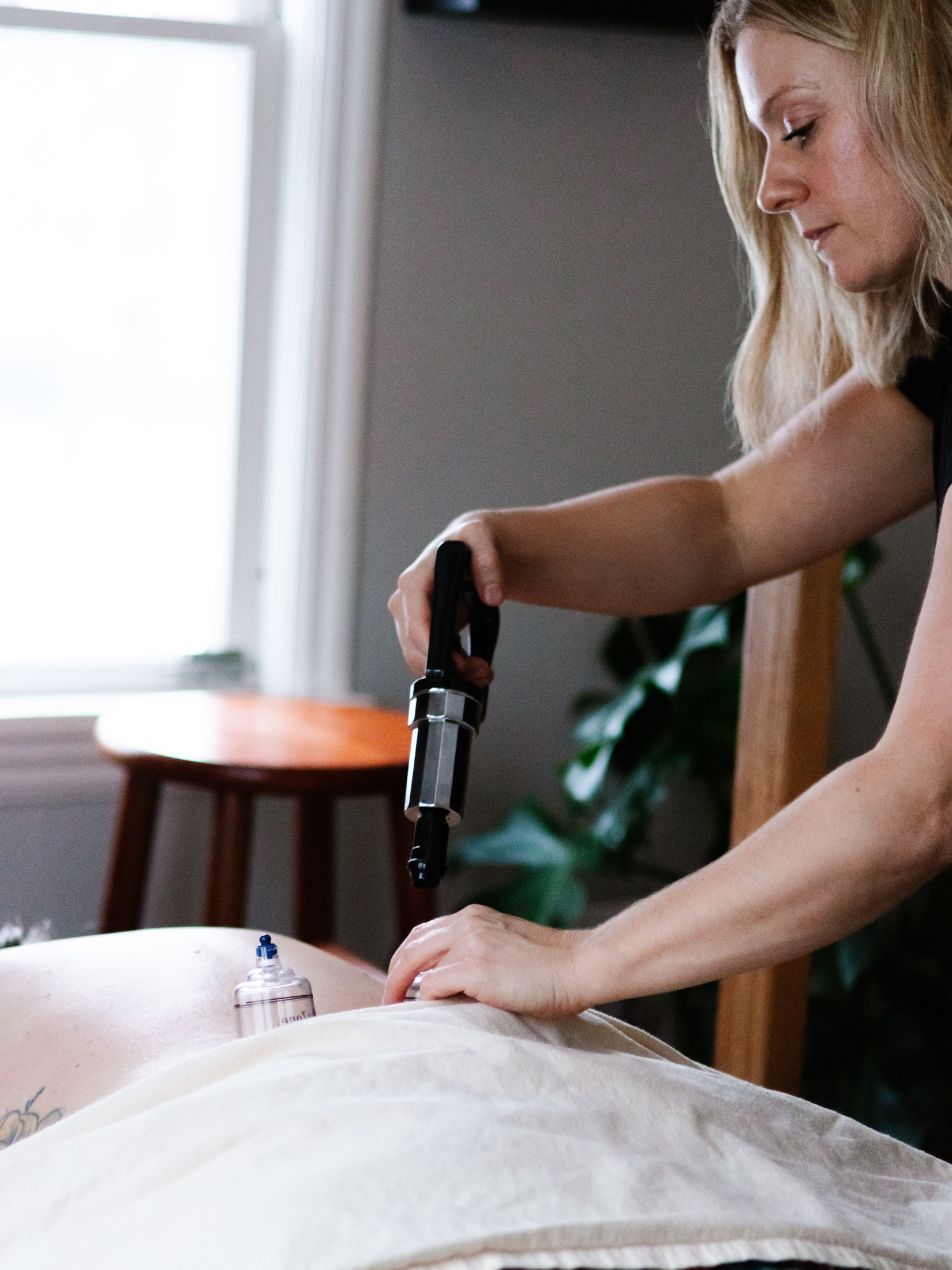 A woman performs a massage therapy with a tool on a person lying face down, with an IV drip nearby, in a room with a window and green plant.