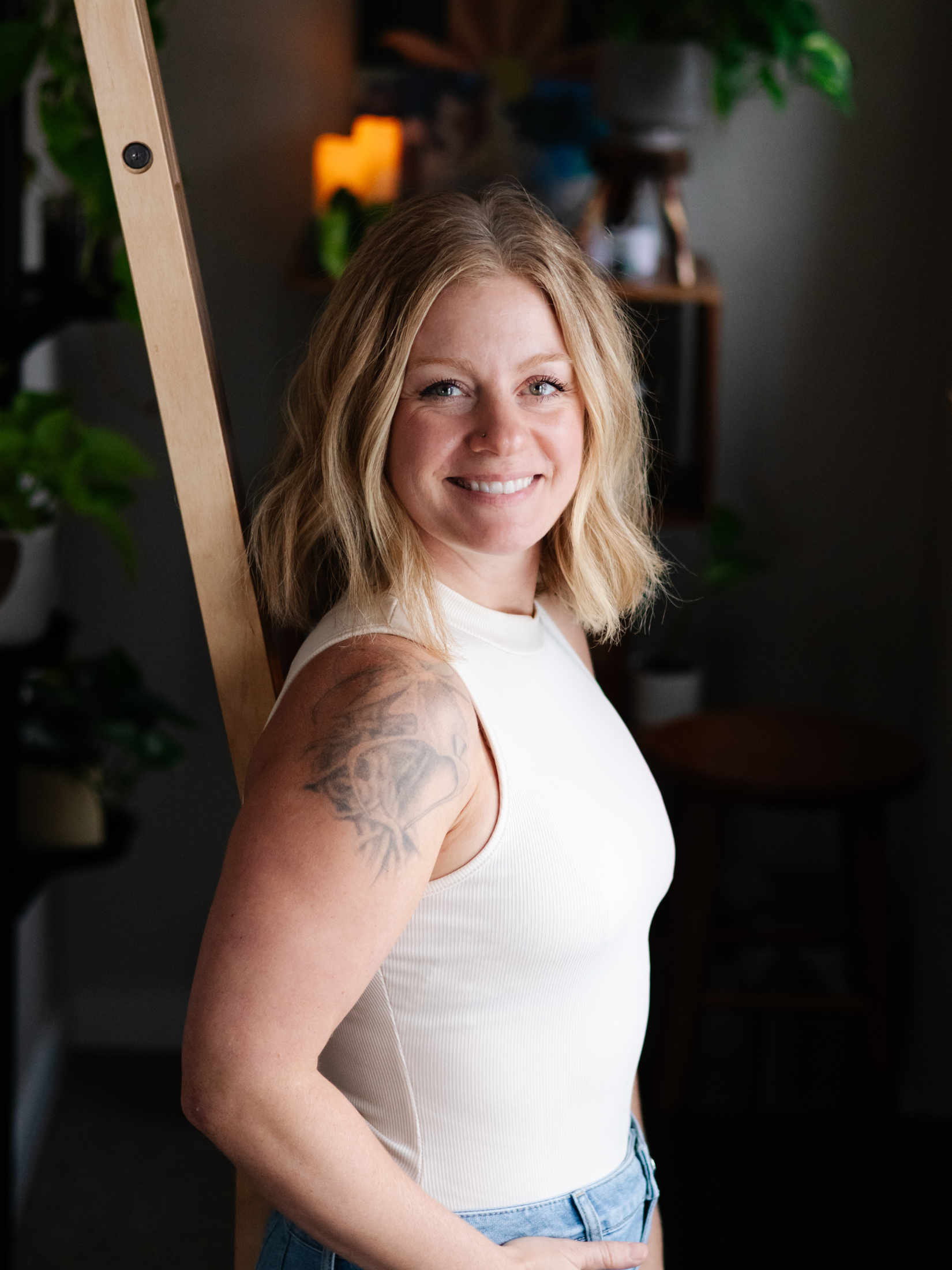 A woman with blonde hair, smiling, standing indoors near an easel, wearing a white sleeveless top and jeans, with plants and shelves in the background.